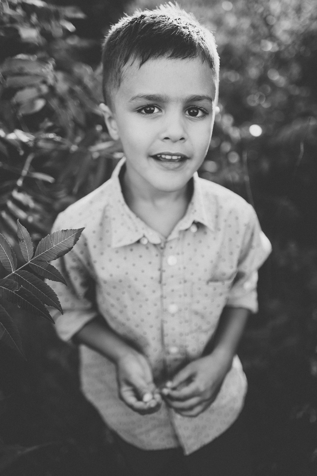 Black and white portrait of young child in polka dot shirt smiling against blurred outdoor background.