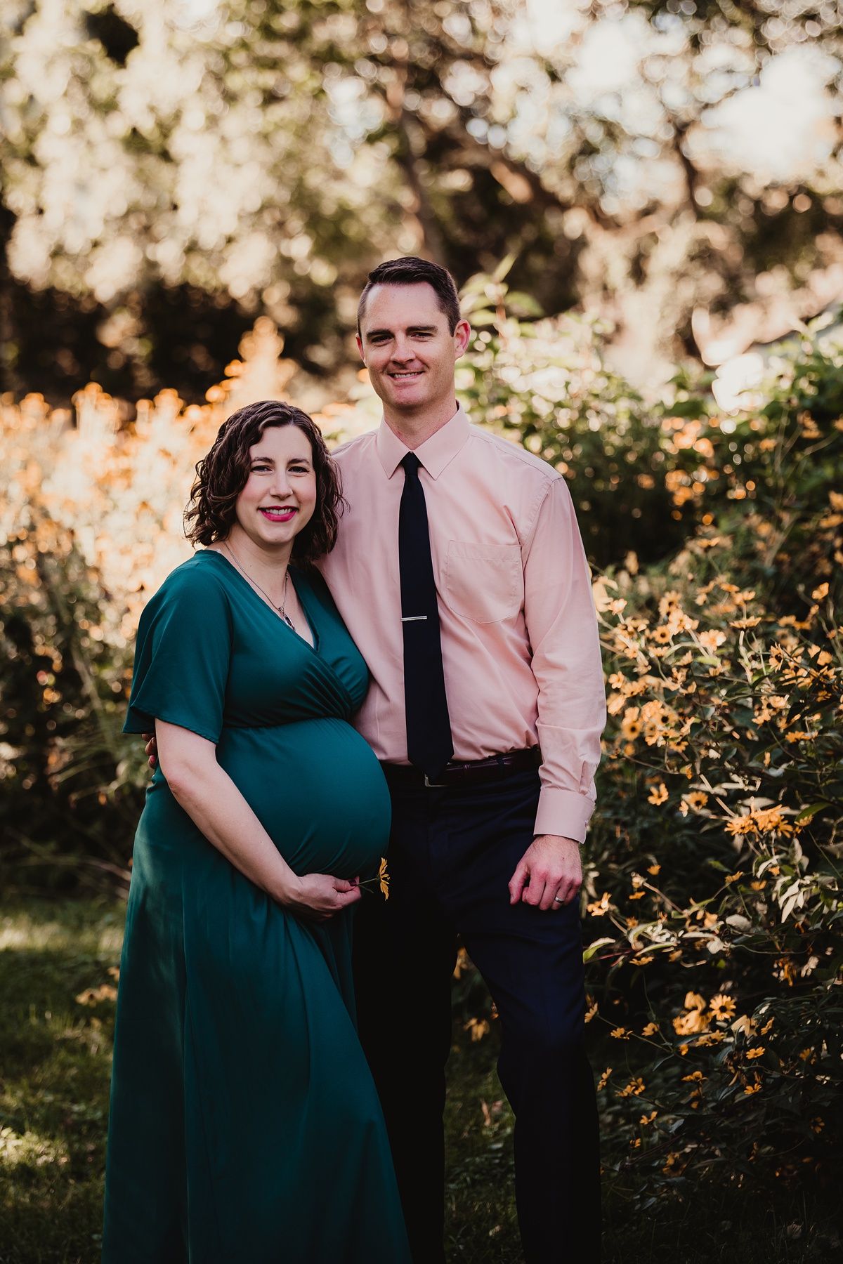 Maternity photo of couple in teal dress and pink shirt standing among yellow flowers at sunset.