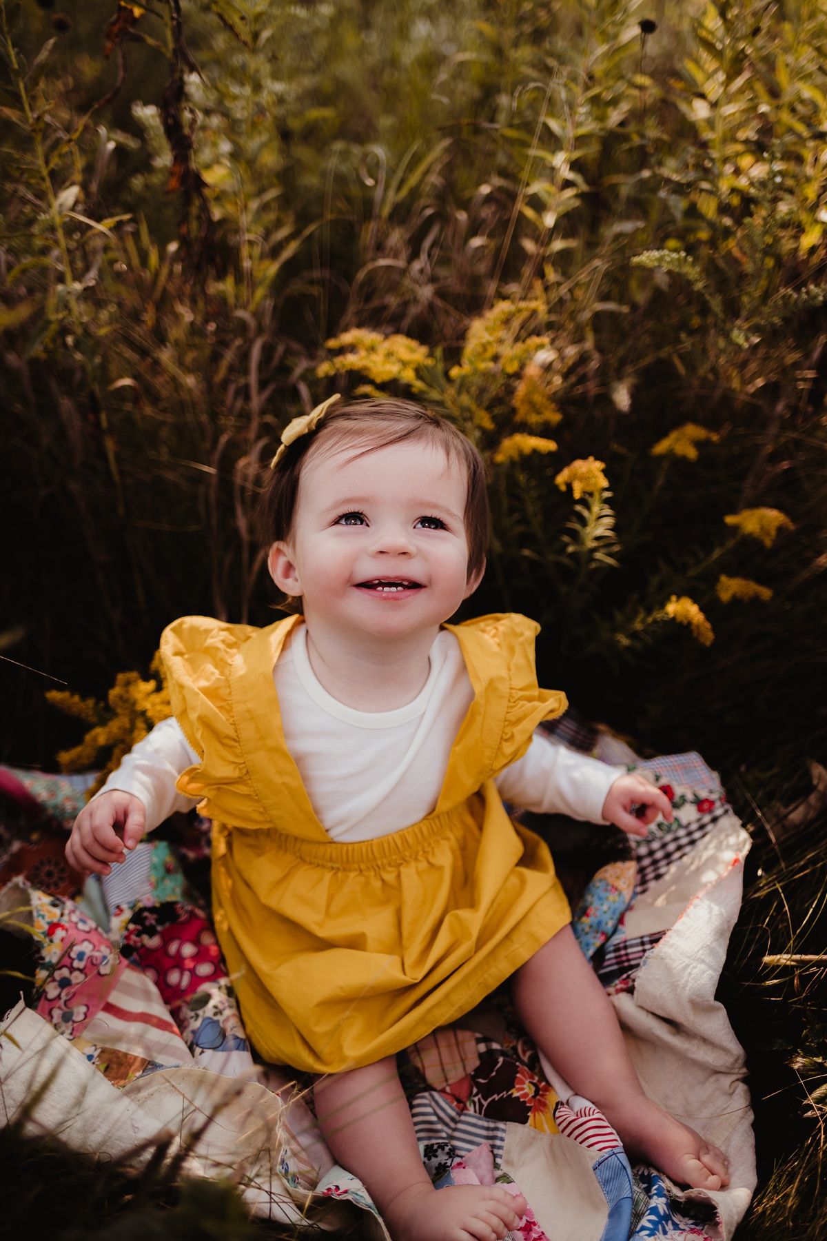 A toddler in a bright yellow dress sits on a floral blanket surrounded by golden wildflowers.
