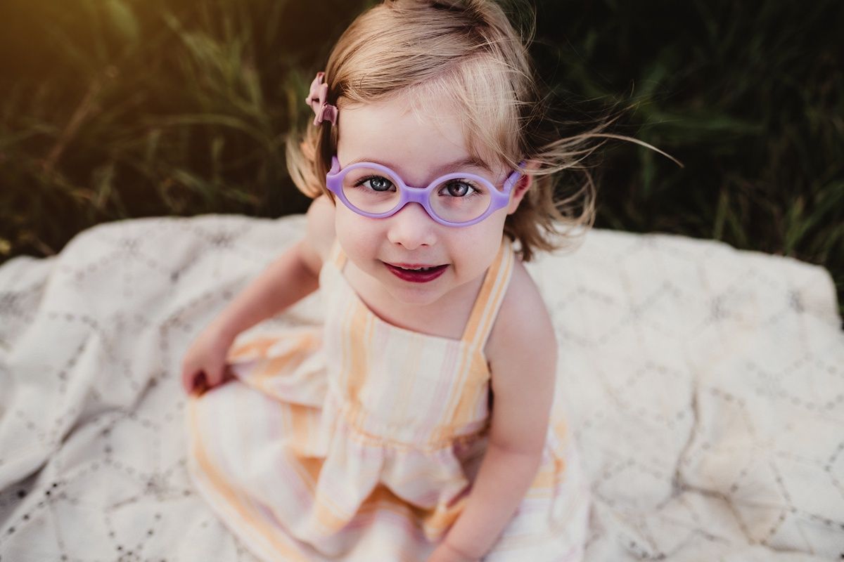 A smiling young child wearing purple glasses sits on a light colored blanket outdoors.