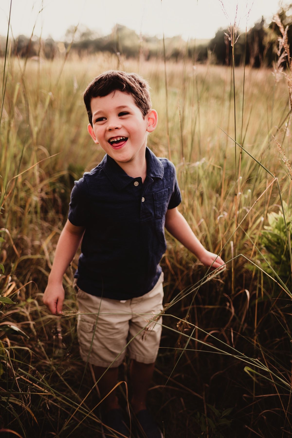 Candid portrait of a child in a navy polo shirt laughing while standing in tall grass at sunset.
