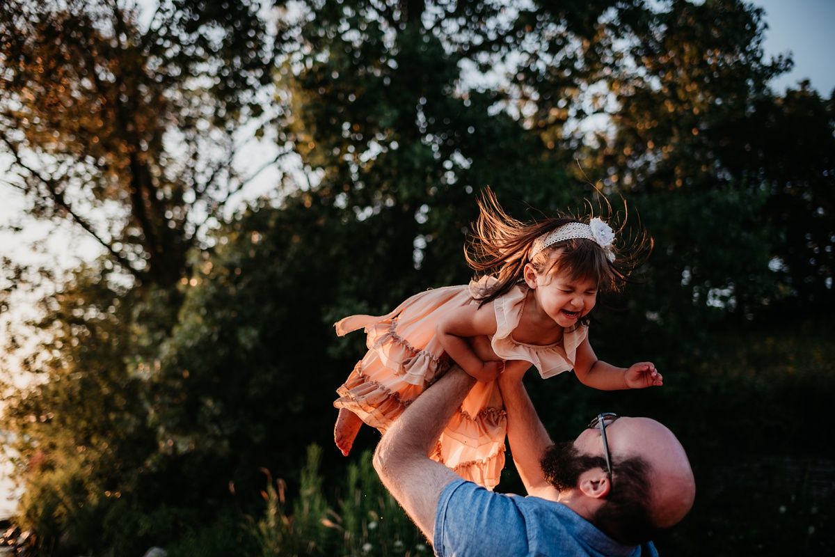 Playful sunset moment at a lakeside as father lifts child high in the air against trees and golden light.