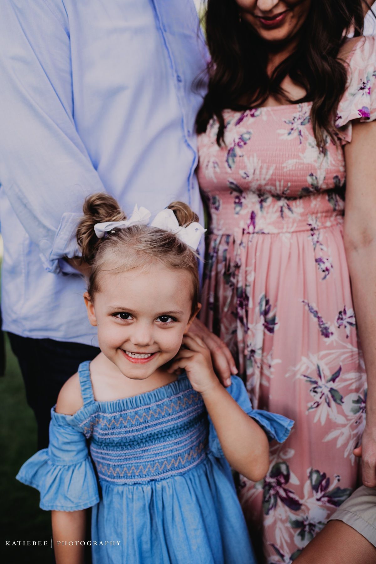 A small child in a blue smocked dress stands with adults wearing a floral pink dress and blue shirt.