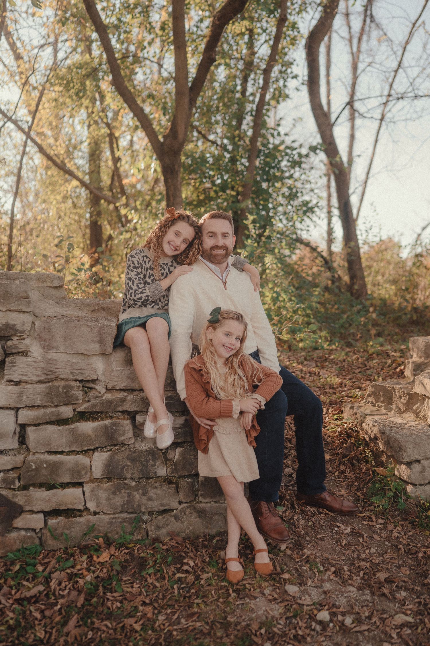 Family group poses on rustic stone wall with fallen leaves and autumn trees in background.
