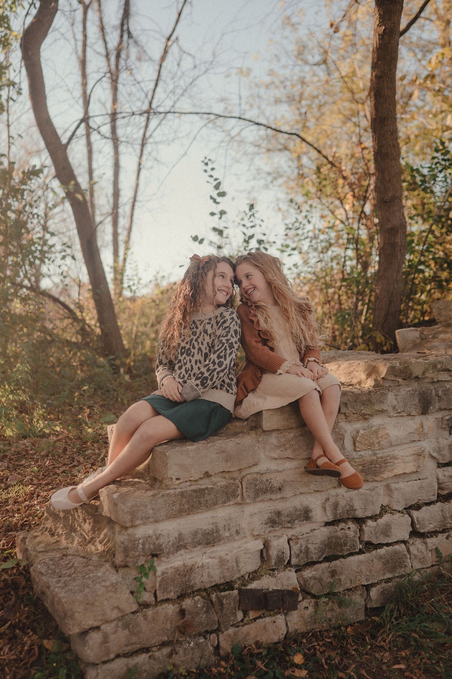Two friends share a candid moment while sitting on a stone wall during golden hour in an autumn forest setting.
