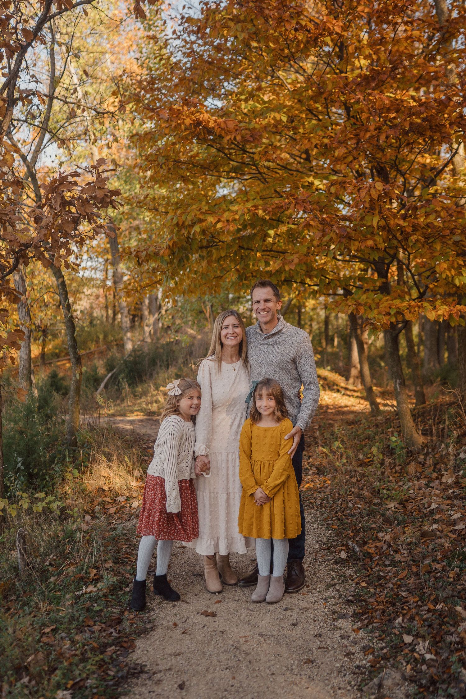 A family poses together on a wooded path surrounded by vibrant autumn foliage in warm golden light.