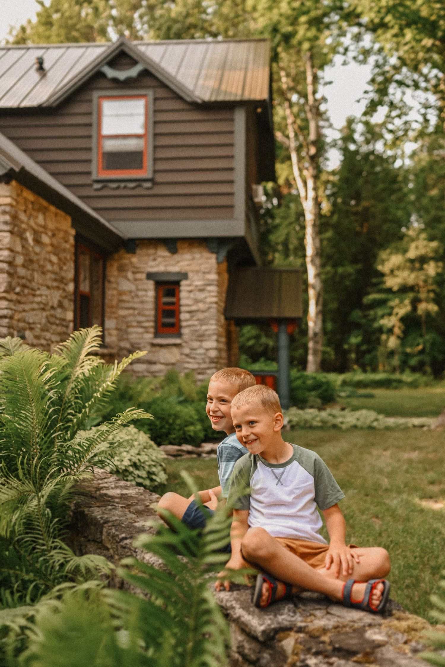 Two children sitting on stone wall near rustic house with ferns in foreground.