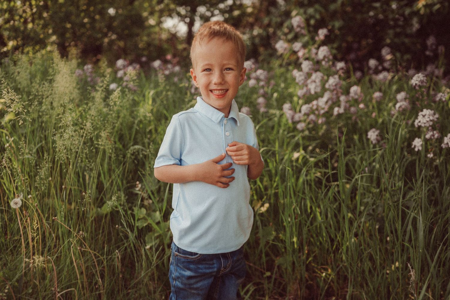 A child in a light blue polo shirt stands joyfully among tall grass and flowering plants.