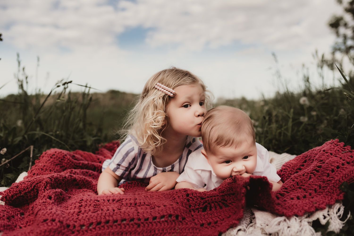 Two children resting together on a red knit blanket outdoors on a sunny day.