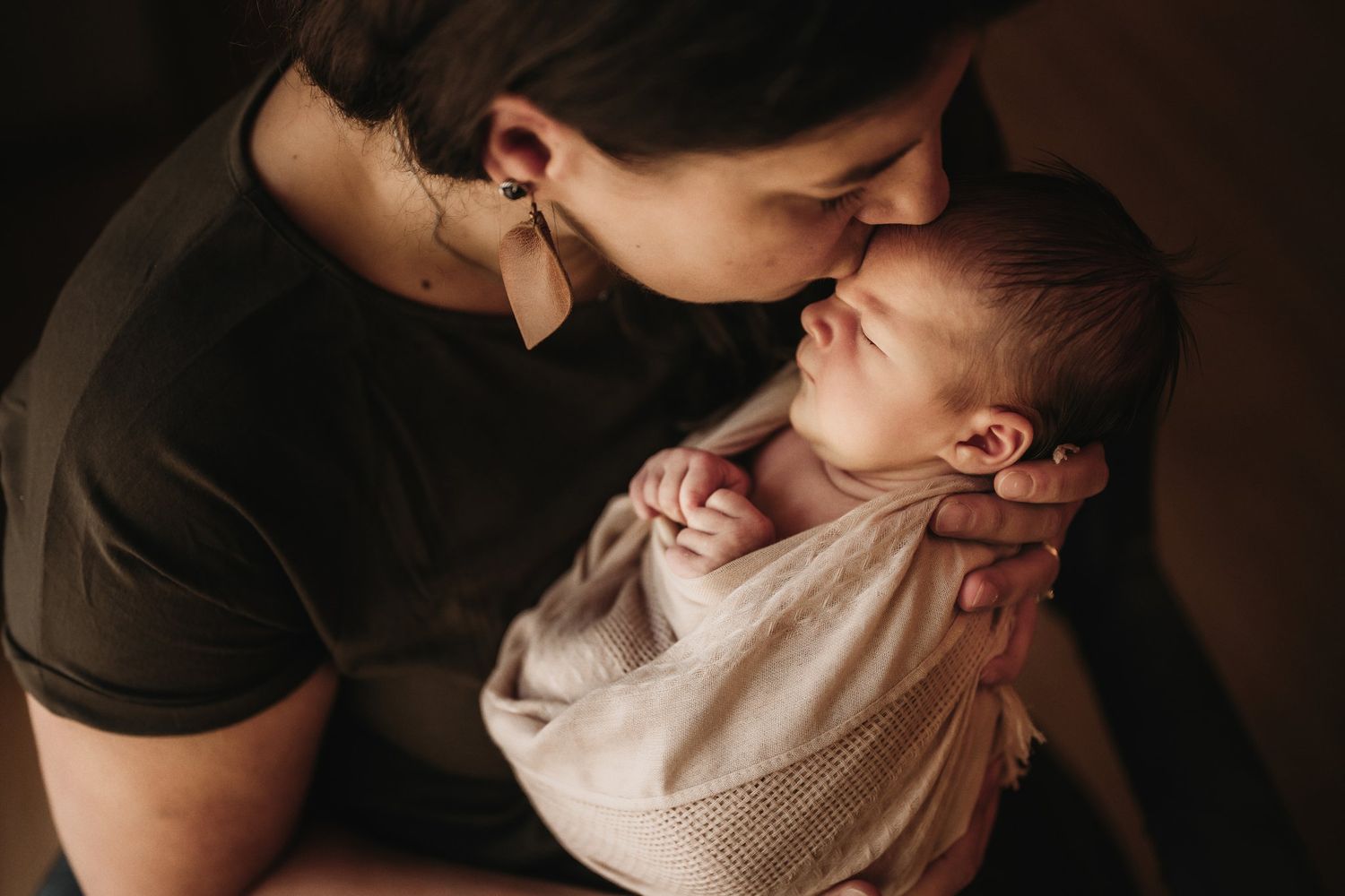 A mother in black gently kisses her swaddled newborn baby in soft lighting.