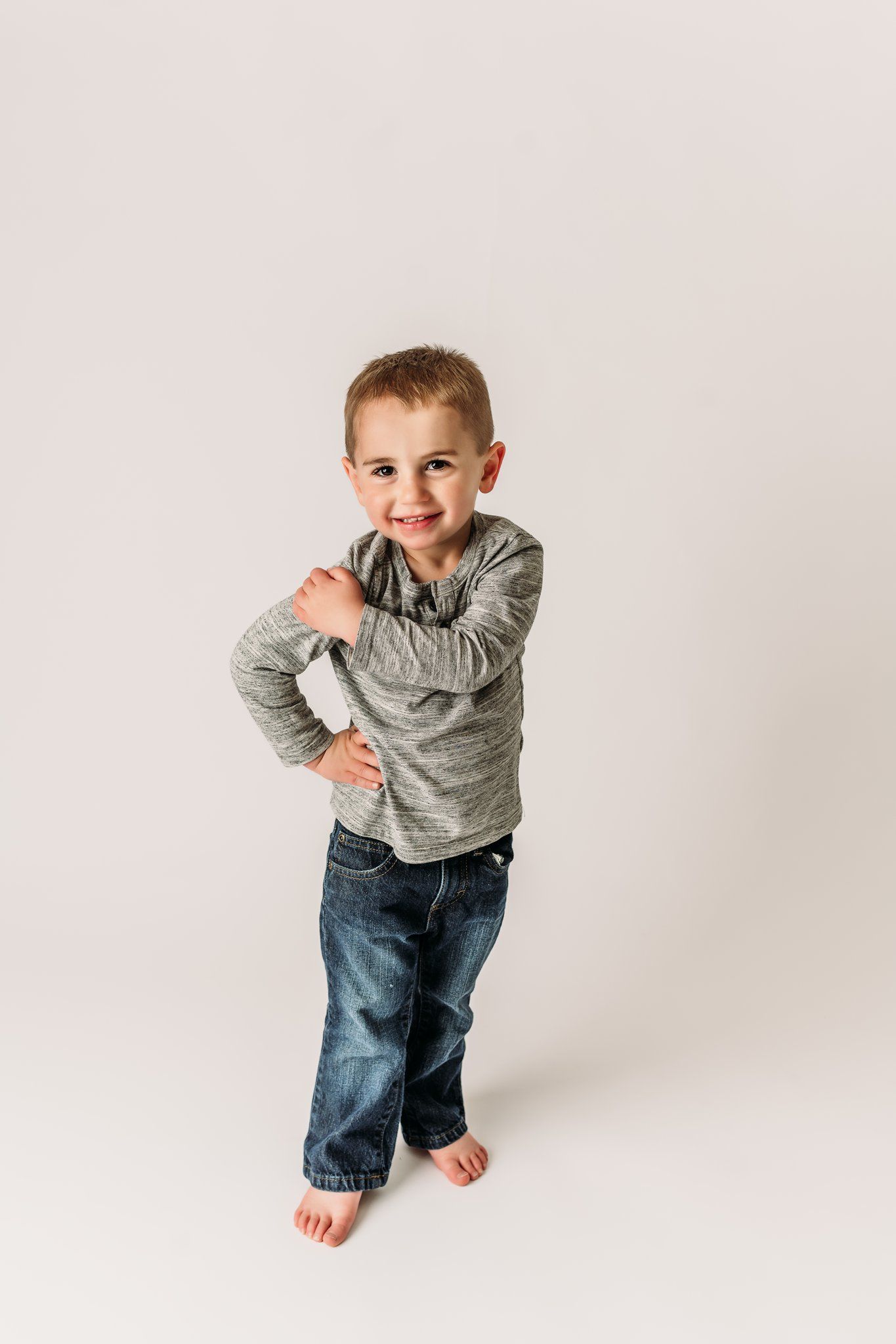 Young child in gray sweater and jeans strikes playful pose against white background.