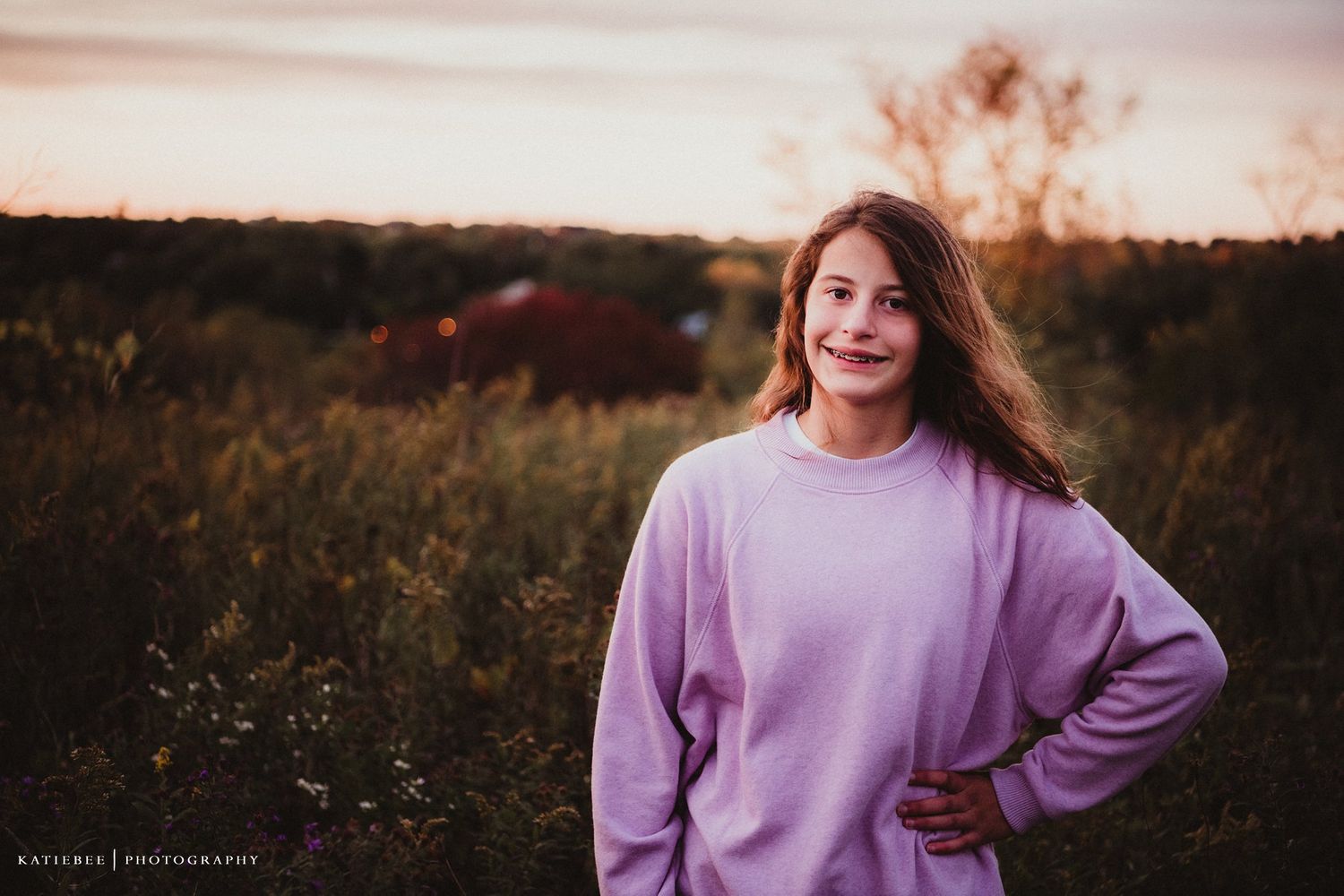 Woman in purple sweater poses confidently in field at sunset.