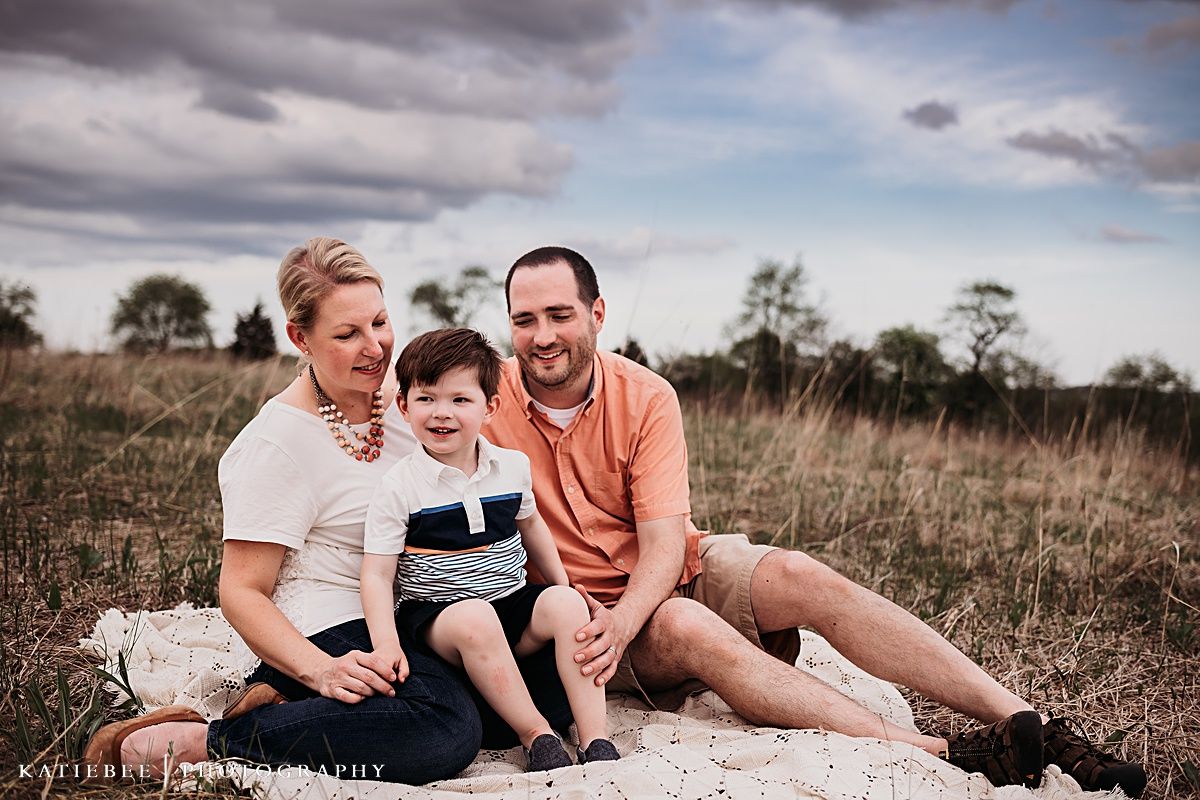 A family poses together on a blanket in a grassy field under dramatic cloudy skies.