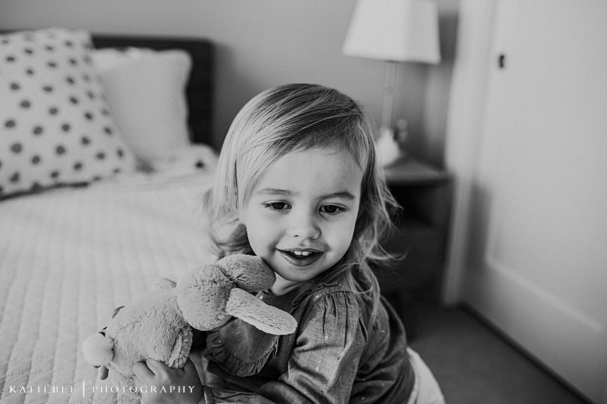 A happy toddler in chambray holding a plush toy sits on a polka dot bedspread.