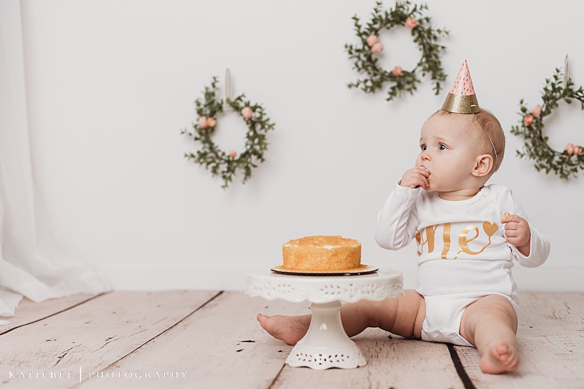Baby in white onesie sits with birthday cake on white pedestal against wall with decorative wreaths.