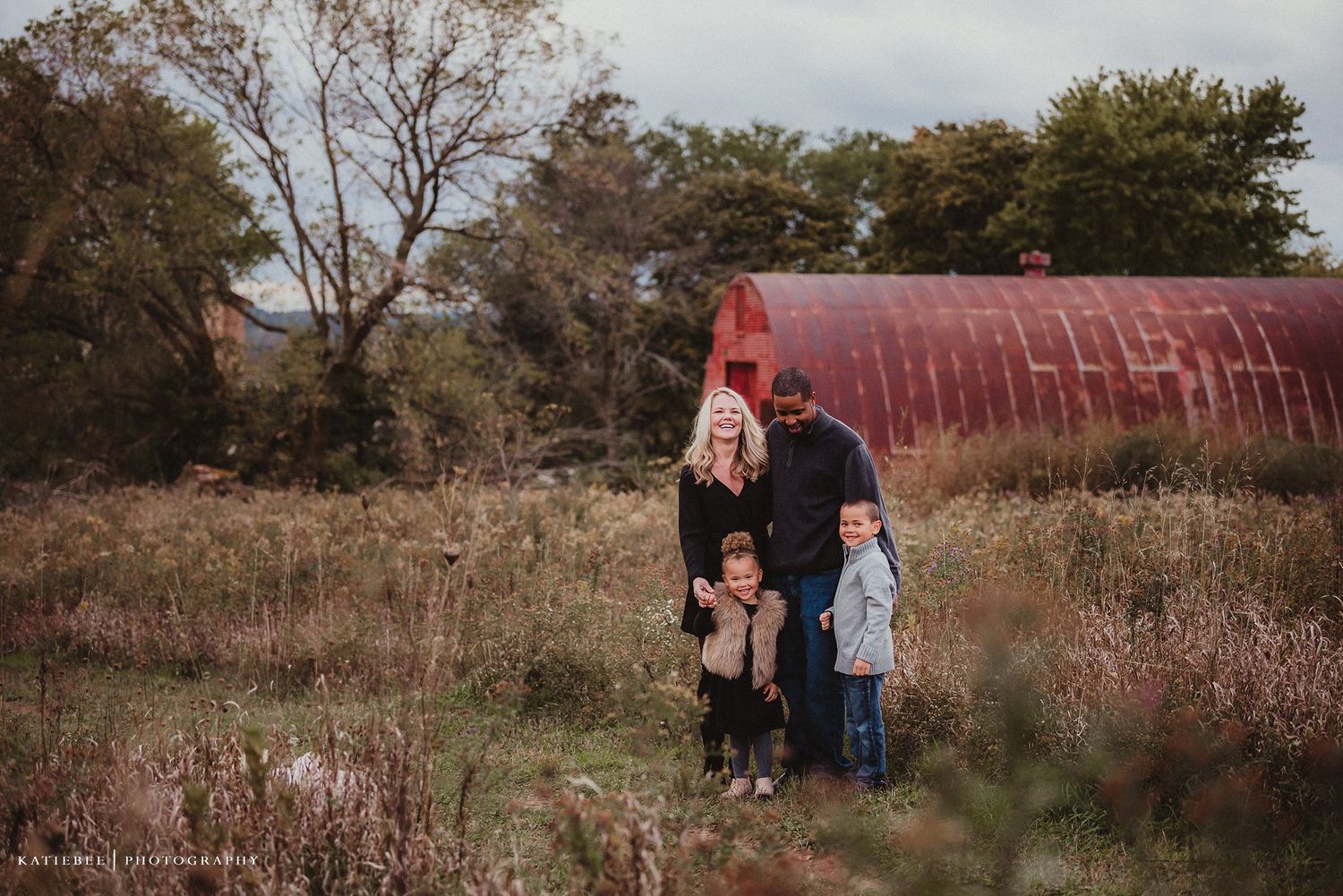 A family standing together in a rustic field with a red barn in the background.