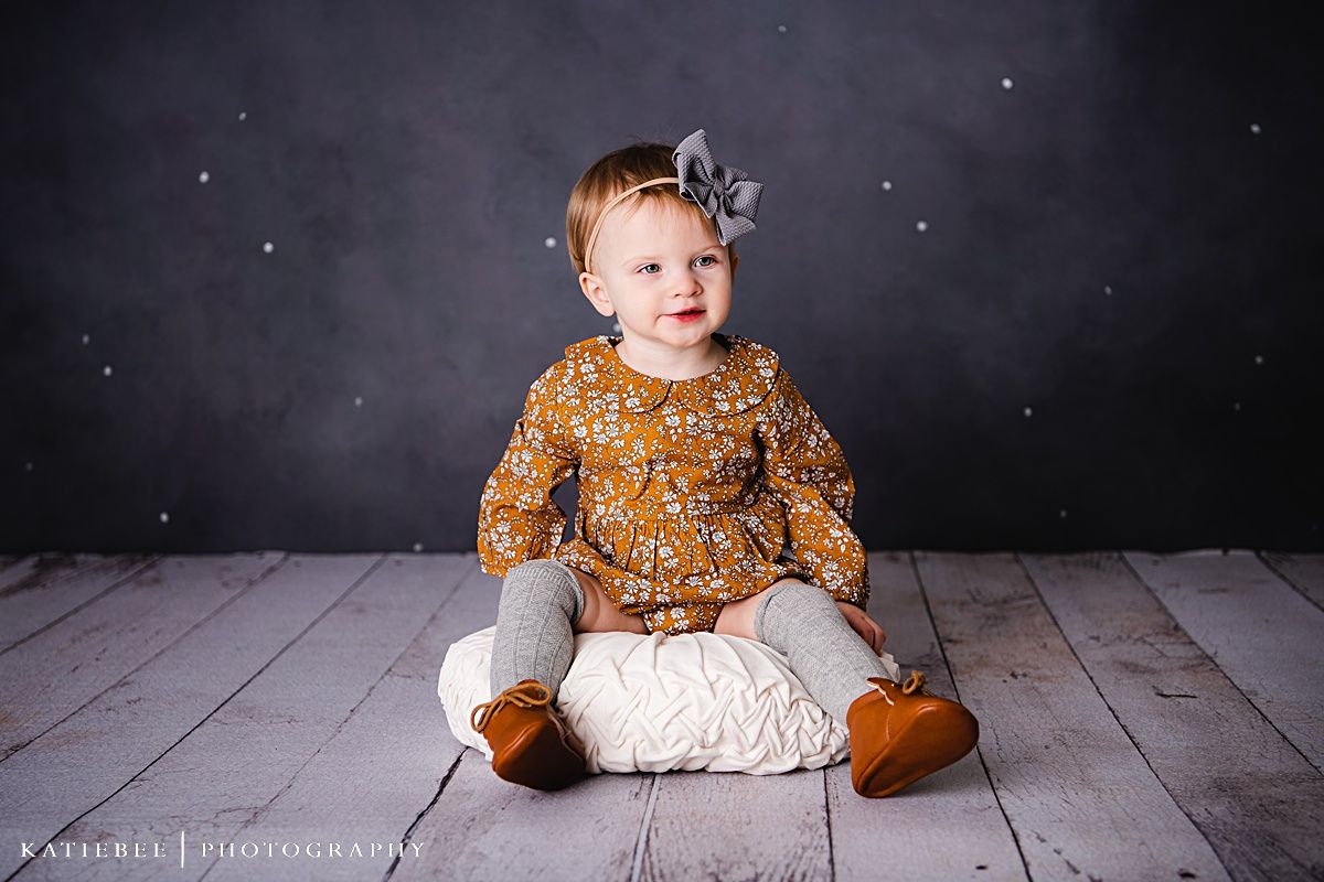 A child in a mustard dress sits on white cushion against wooden floor and dark backdrop.