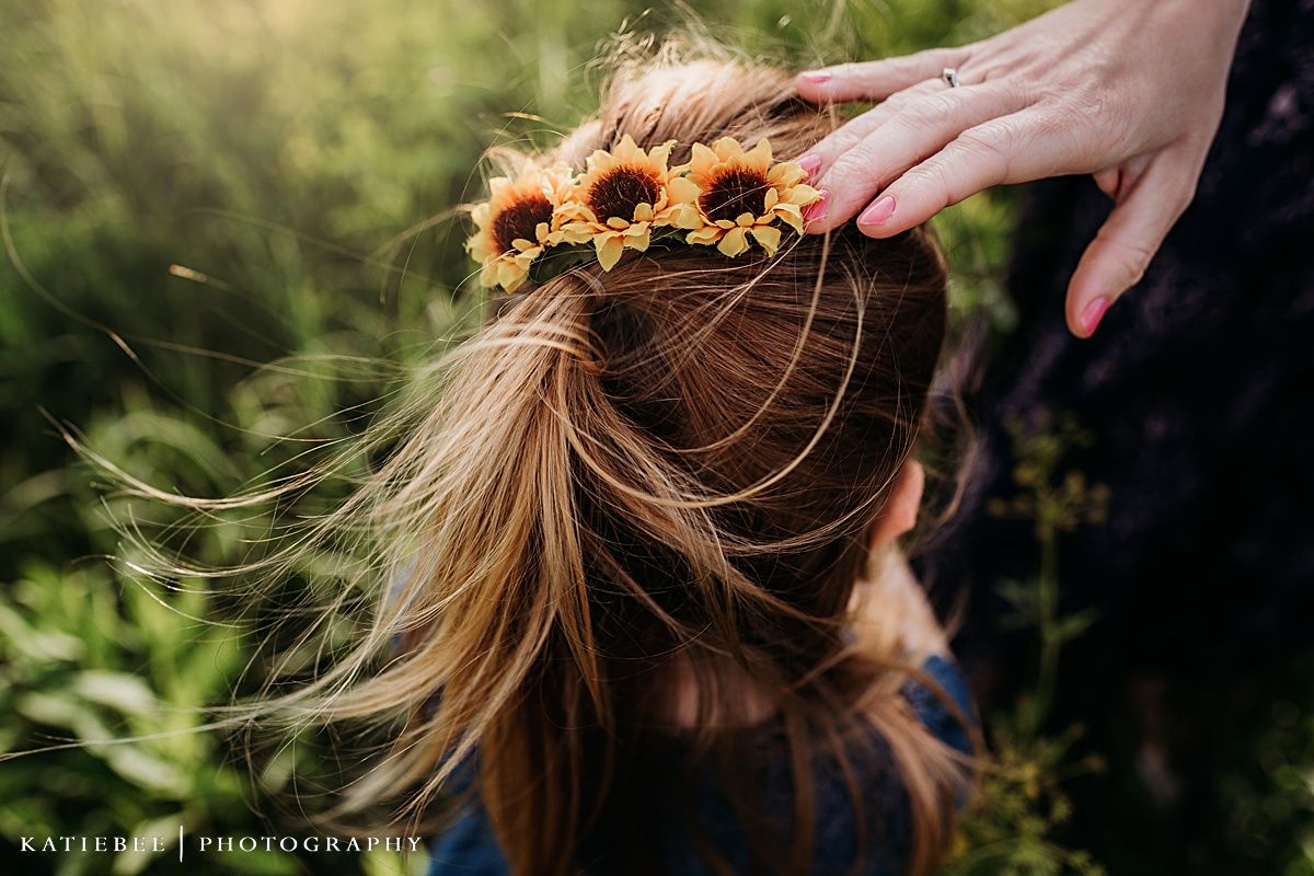 A hand adjusts a delicate crown of sunflowers on a person's hair against a green outdoor background.