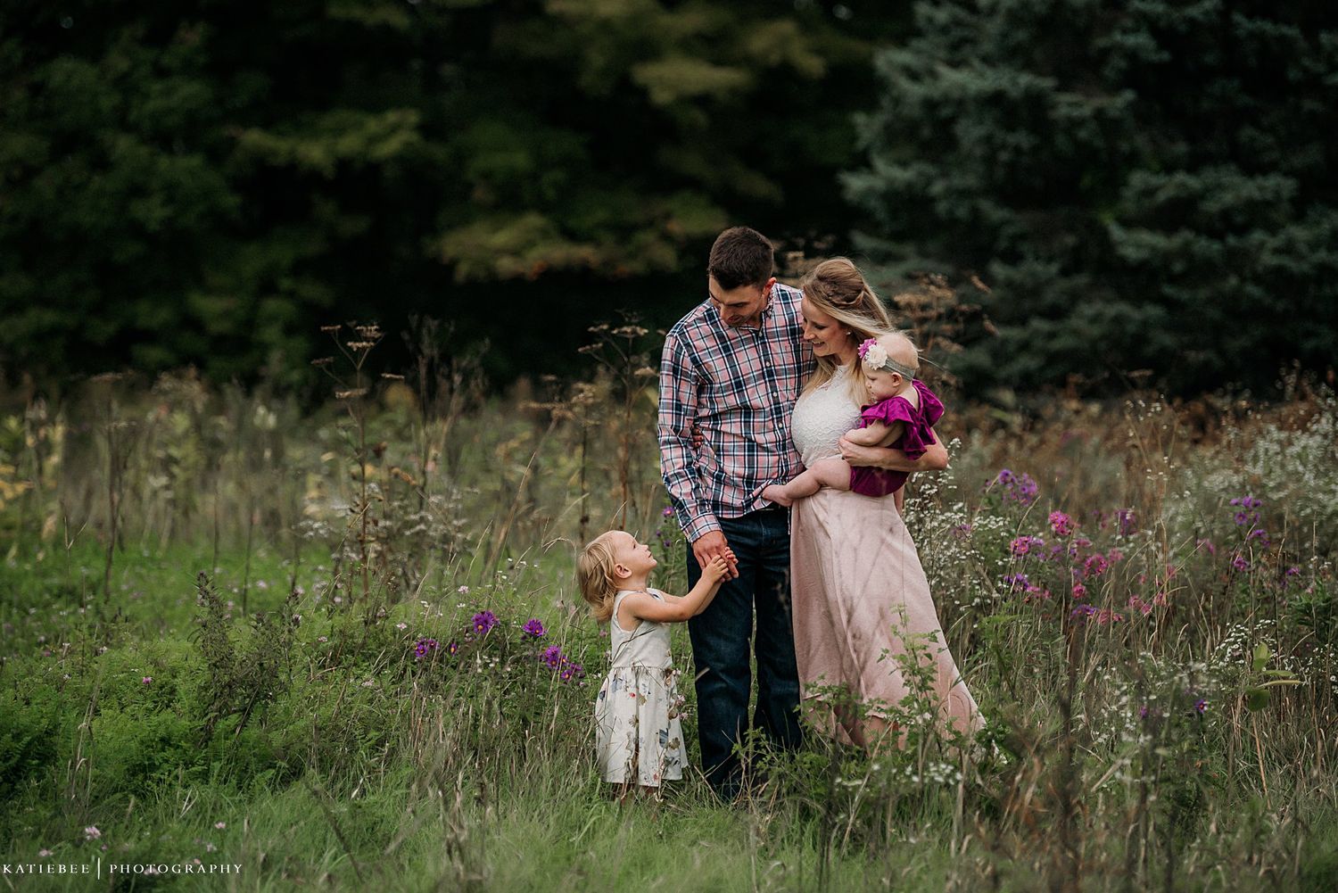 A family shares an intimate moment together in a field with evergreen trees in background.