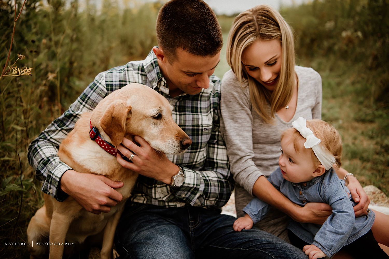 A couple sits outdoors with their golden retriever and baby enjoying a tender family moment together.
