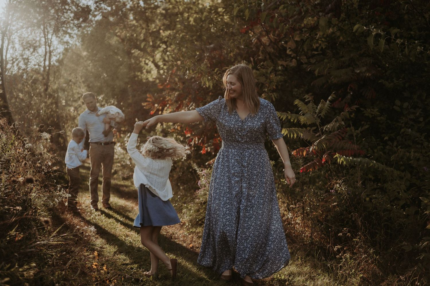 Family members holding hands while walking through a sunlit forest path.