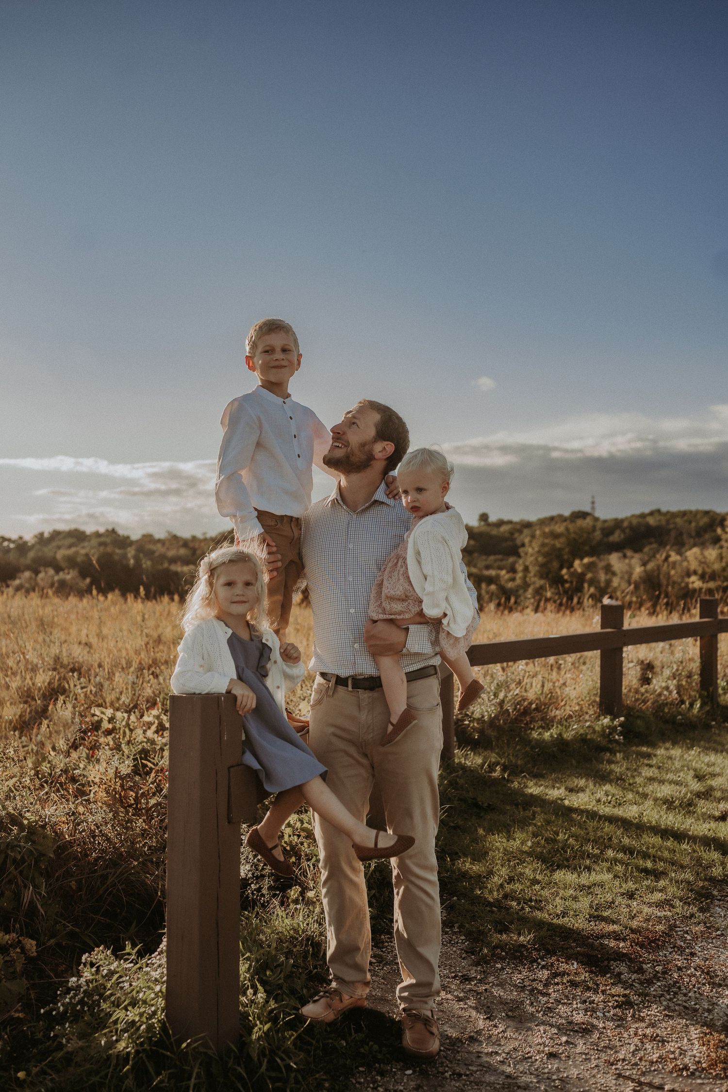 A family portrait captured in golden light against a rustic wooden fence and rolling countryside backdrop.