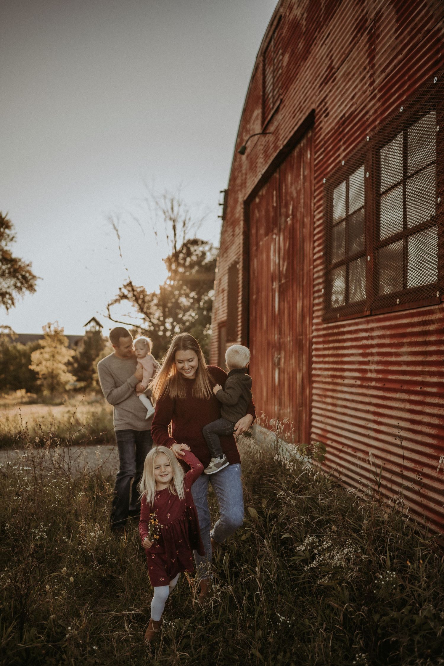 Family photoshoot near a rustic red barn building in golden evening light.