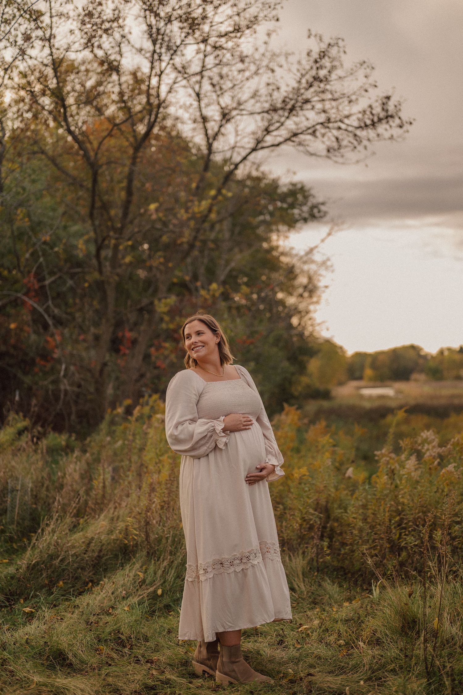 A woman in a flowing beige dress poses in a sunlit autumn meadow surrounded by tall grass and trees.