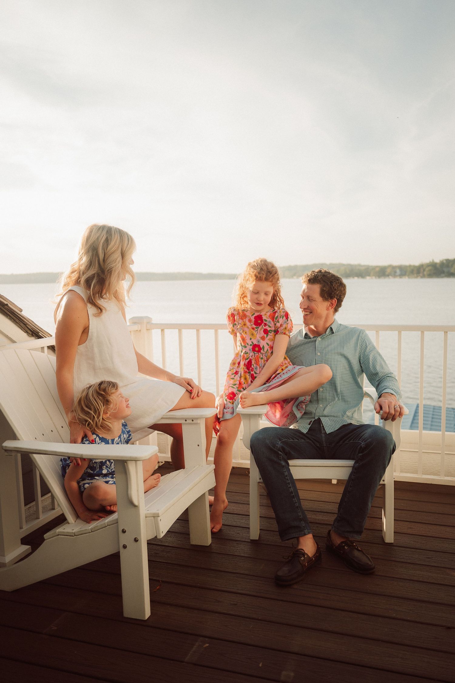 Family members share a relaxed conversation while sitting on white Adirondack chairs on a waterfront deck.