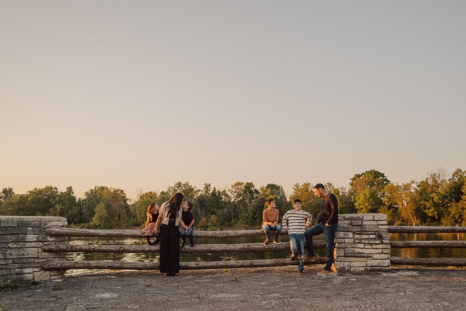 People relaxing on a stone wall overlooking a tranquil lake at dusk with autumn trees in the distance.