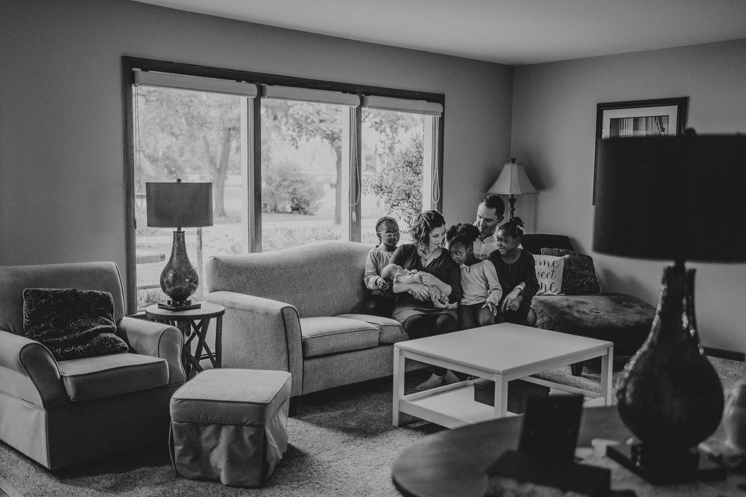 Black and white photo of family gathered on living room sofa near large windows.