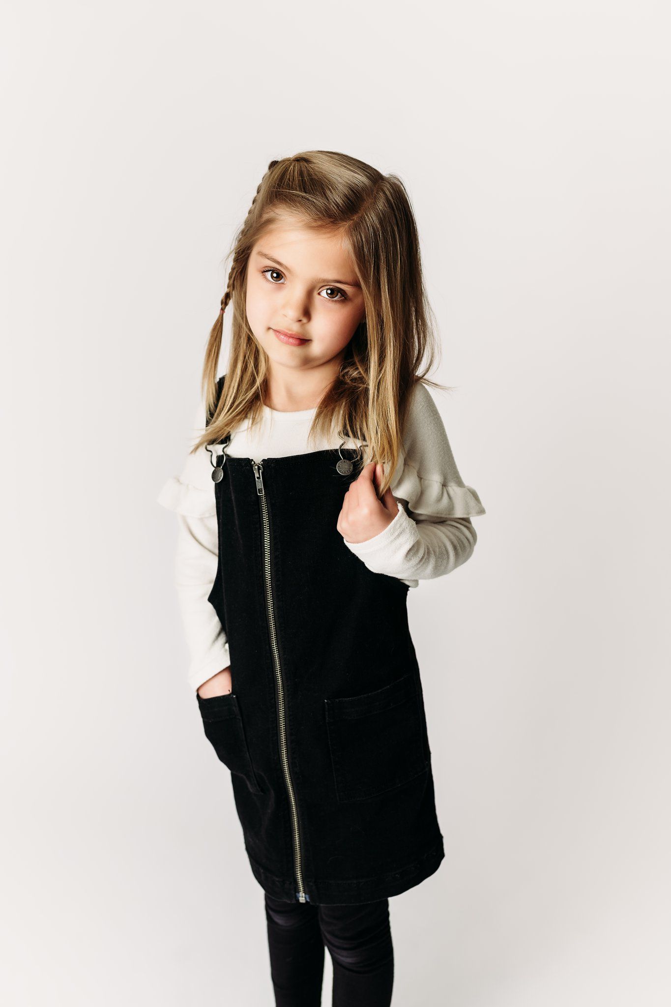 A child wearing a black jumper dress with white long sleeves stands against a white backdrop.