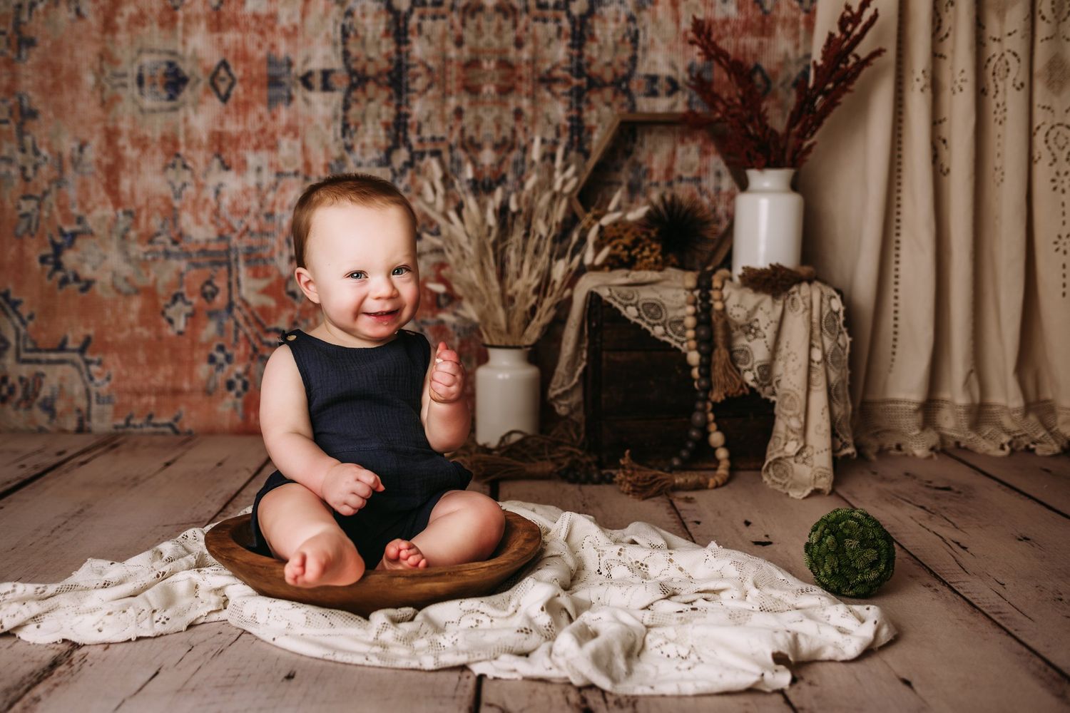 Smiling baby in navy outfit seated in wooden bowl with dried flowers and vintage decor.
