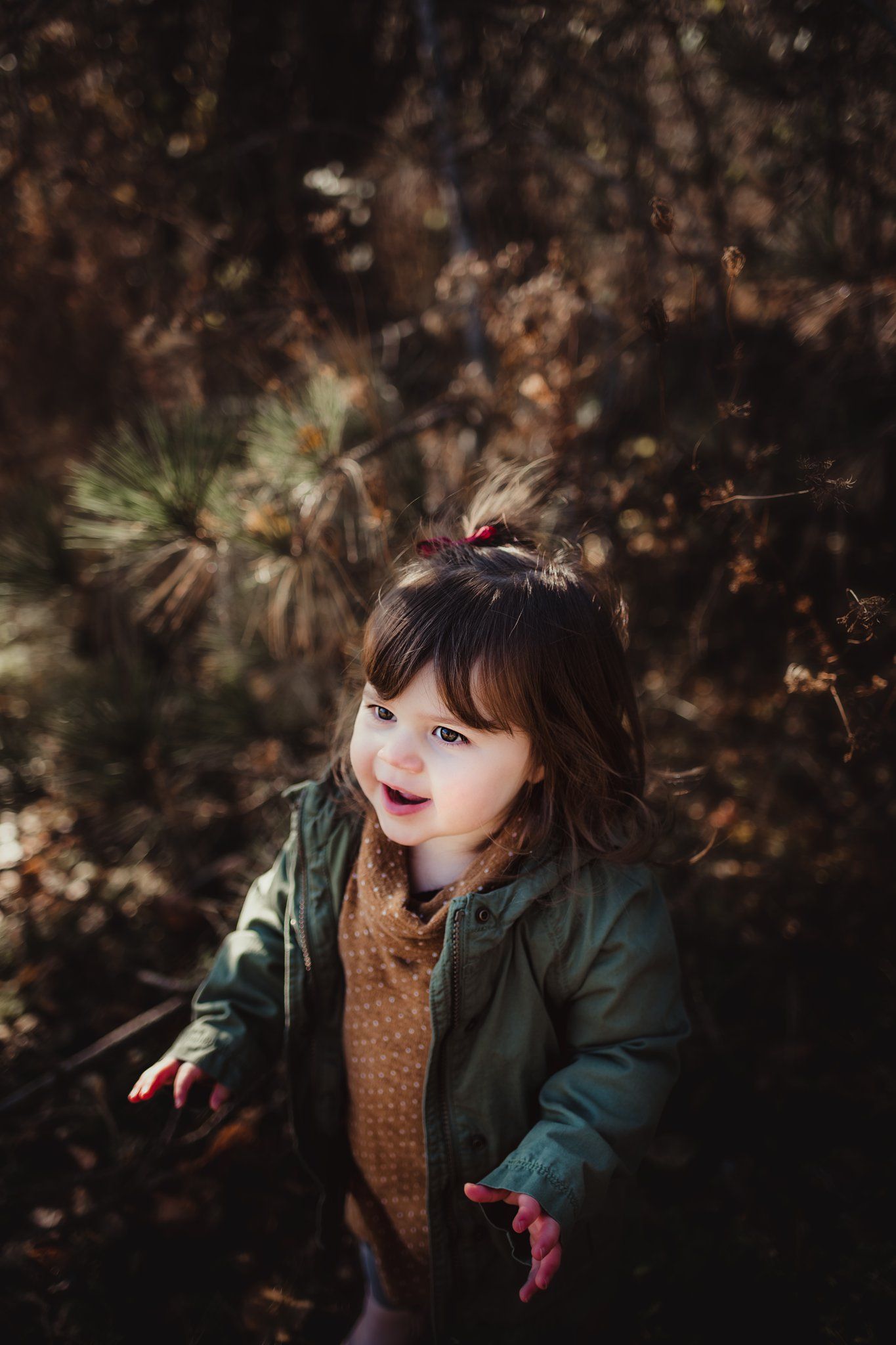 A child wearing a green jacket and brown sweater smiles playfully in an autumn forest setting.