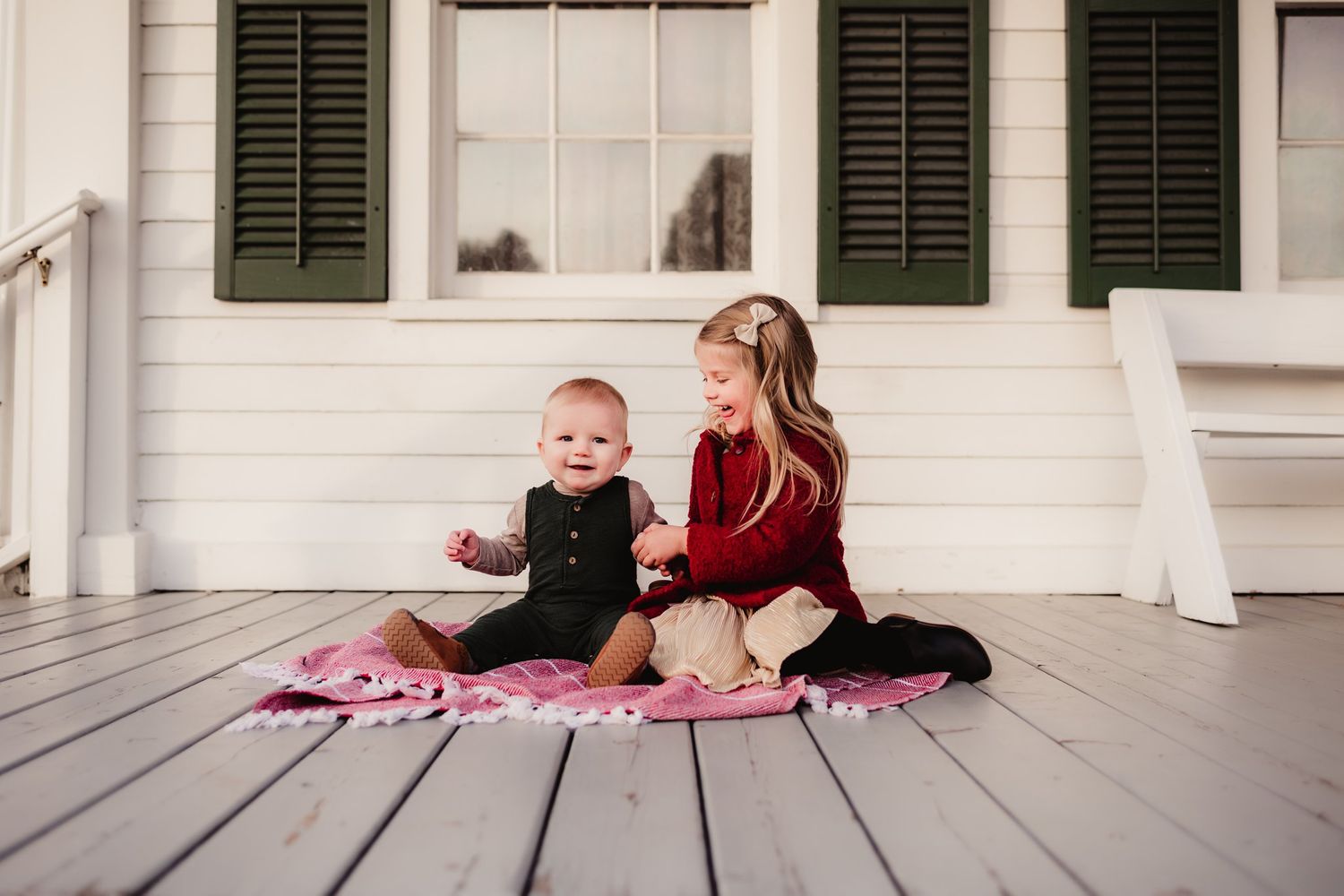 Two children sitting together on pink blanket on white porch.