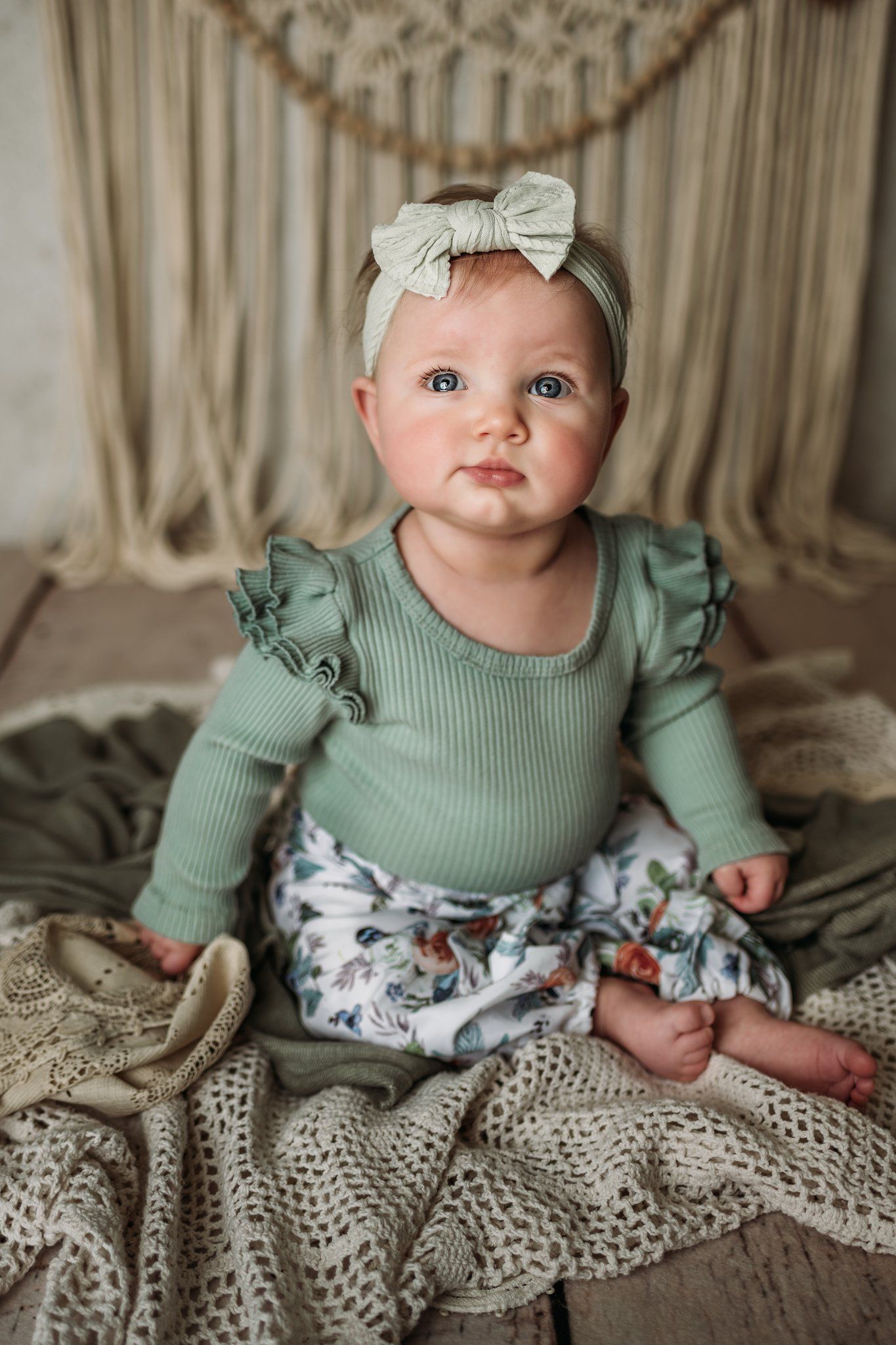 Baby wearing sage green ruffled outfit and white bow headband sits on knit blanket with macrame backdrop.