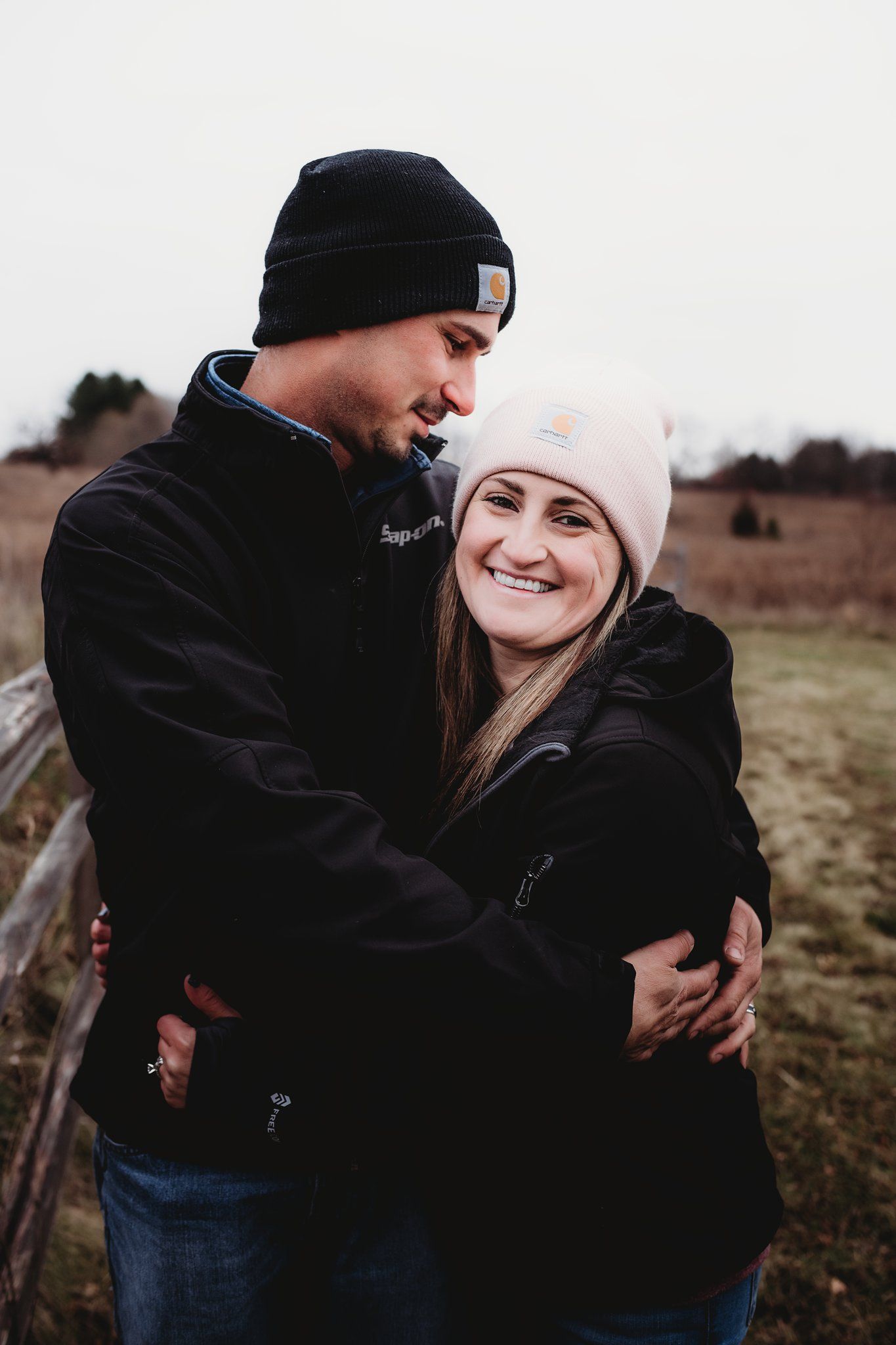 A couple in winter beanies embrace and smile while standing in an open field on an overcast day.