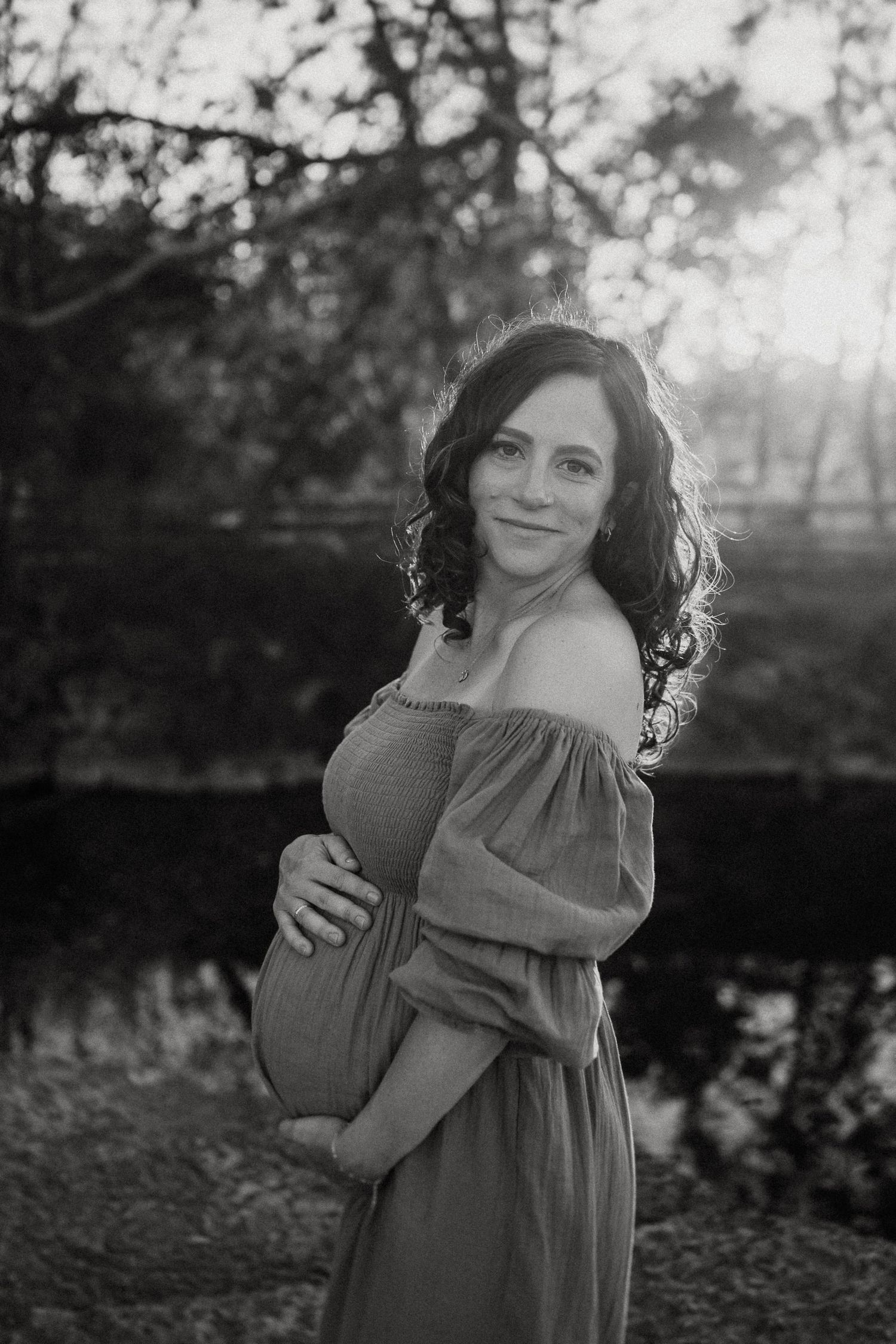 A maternity photo in black and white shows a woman in an off-shoulder dress standing in nature during golden hour.