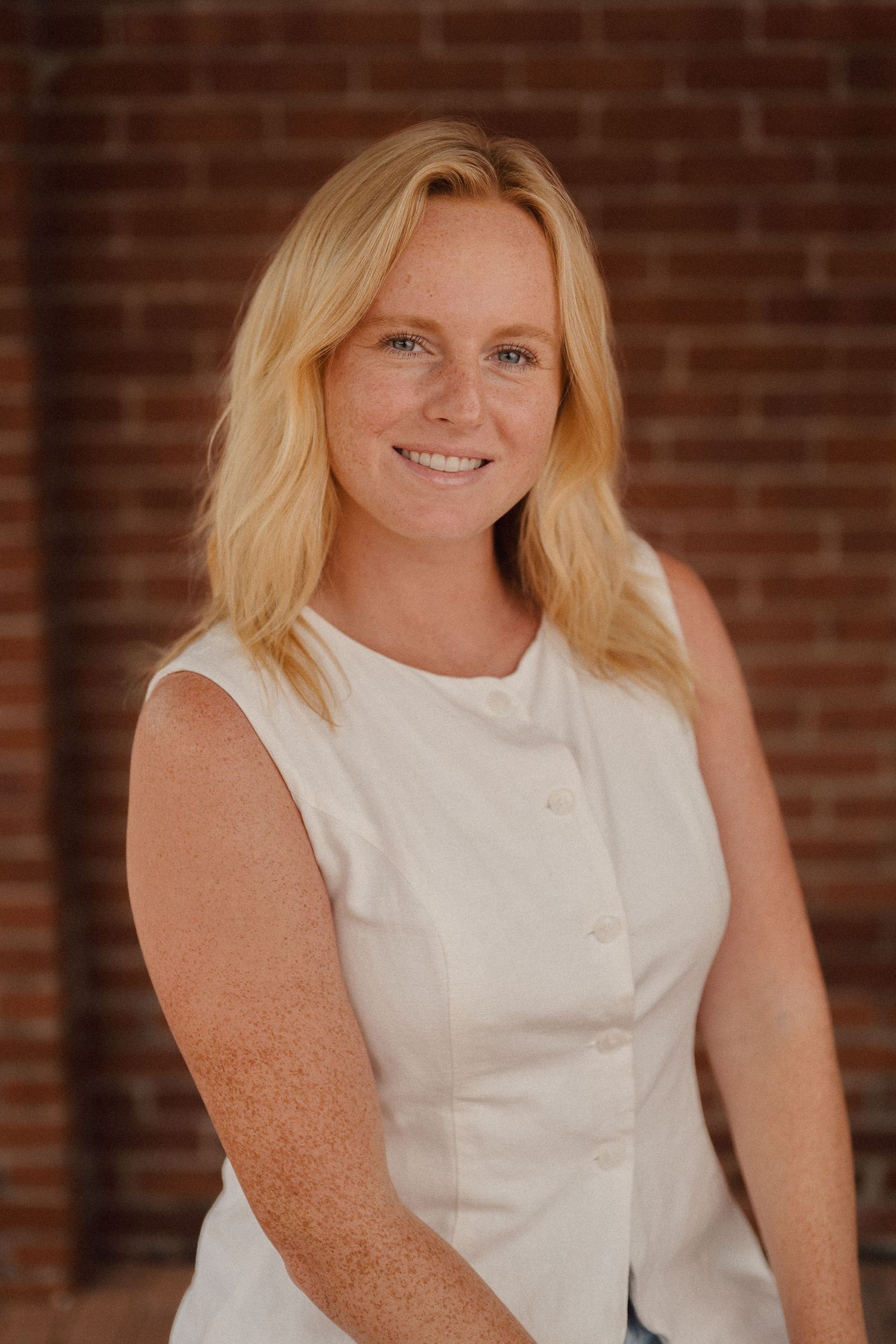 Professional headshot of a person in sleeveless white top against rustic brick wall background.