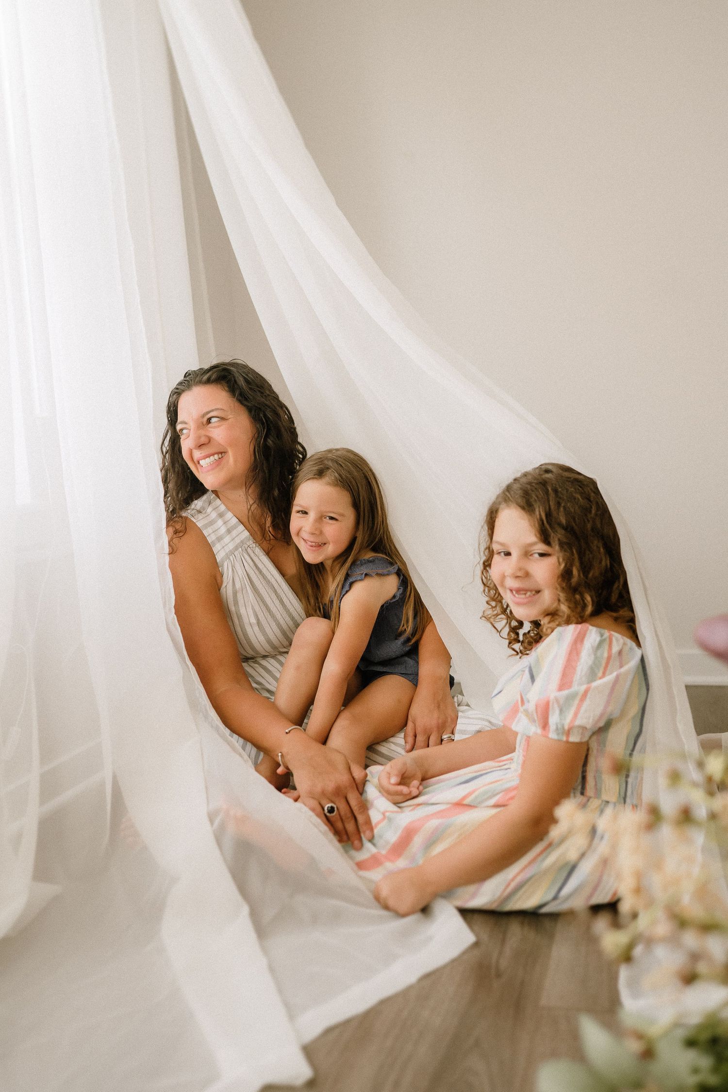Family shares joyful laughter while sitting together against ethereal white drapes in a lifestyle portrait.