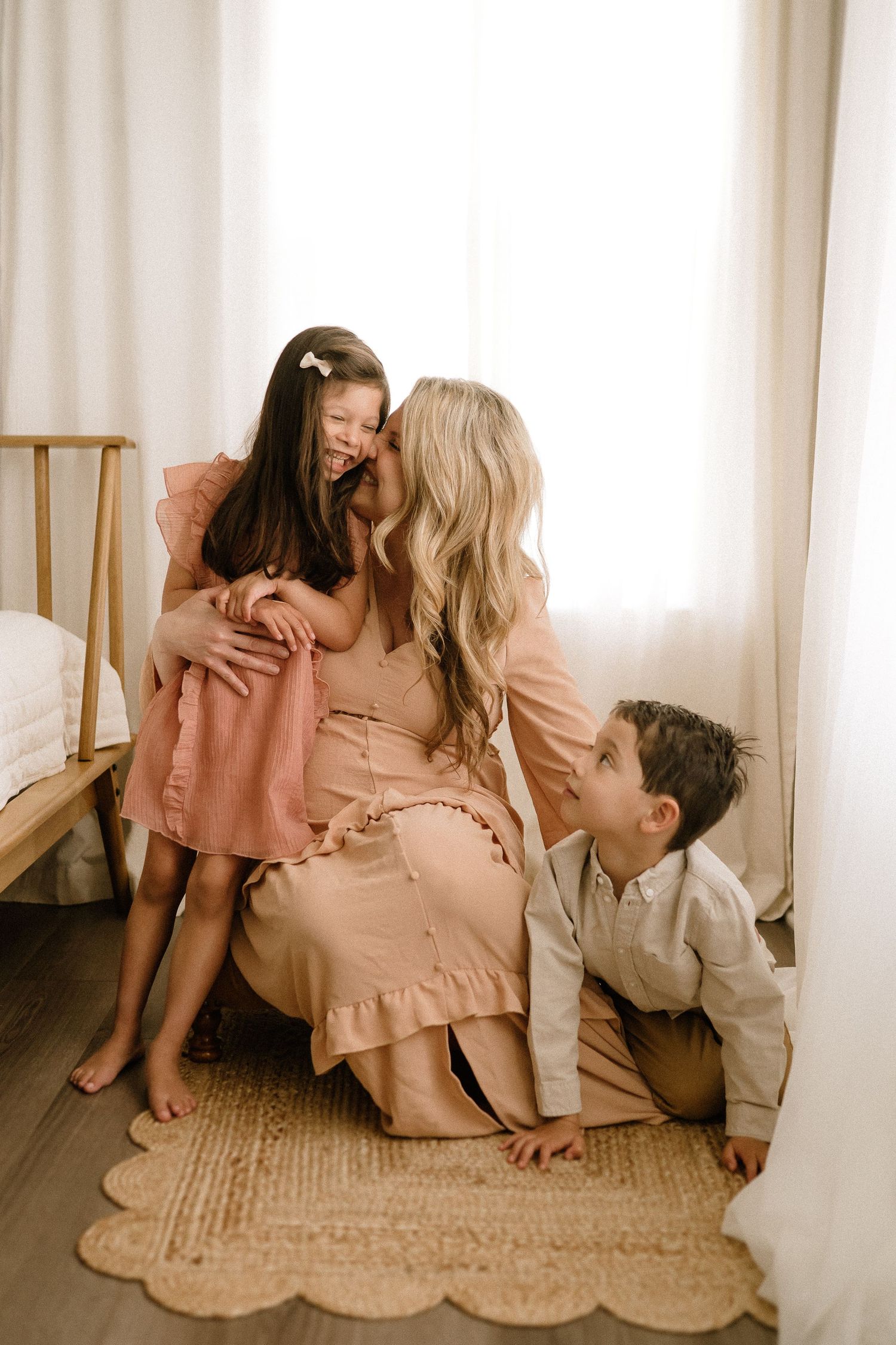 Candid family moment shows mother and children sharing tender interaction on a jute rug in sunlit room.