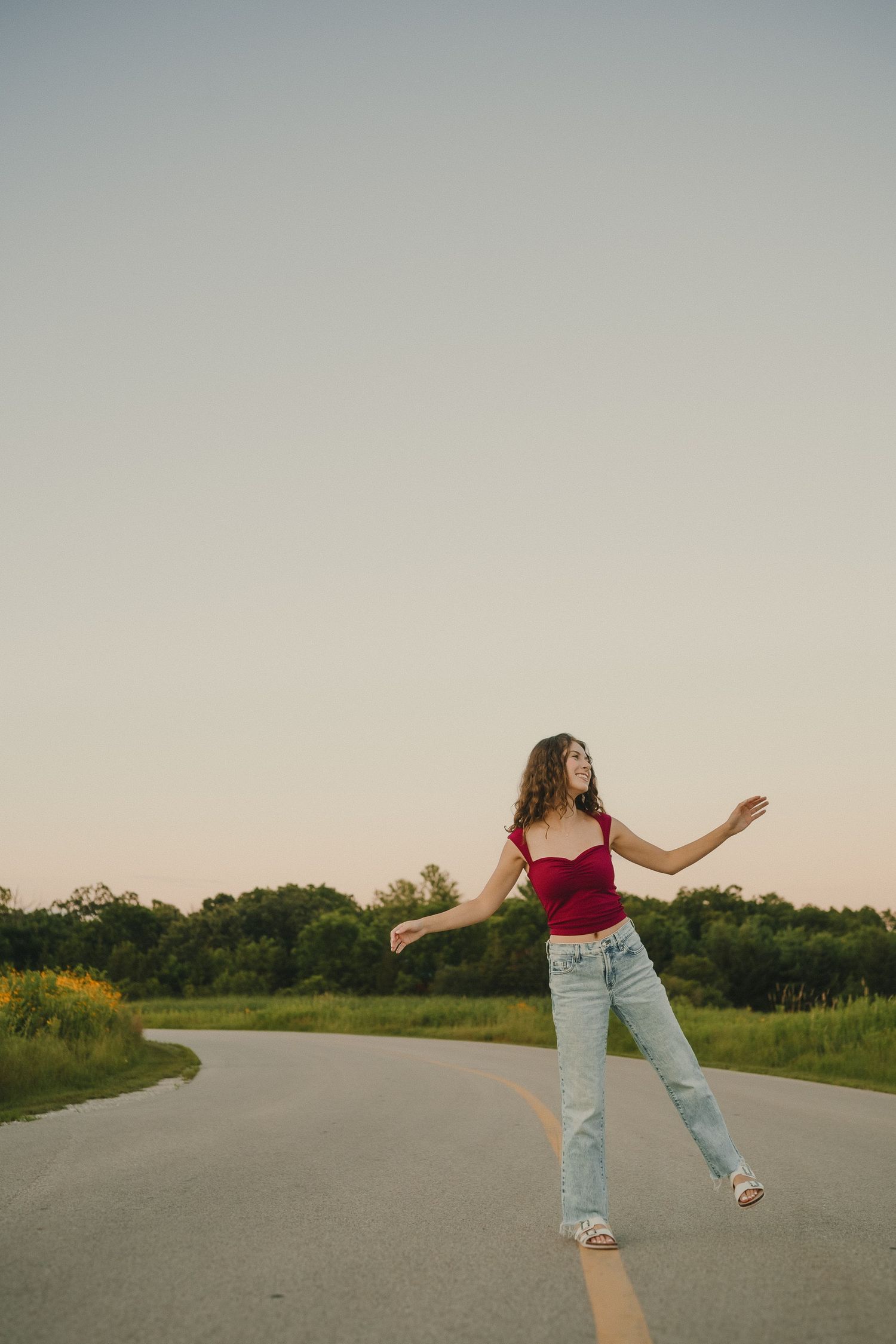 Person wearing red tank top and light jeans dances carefree on an empty rural road during golden hour sunset.