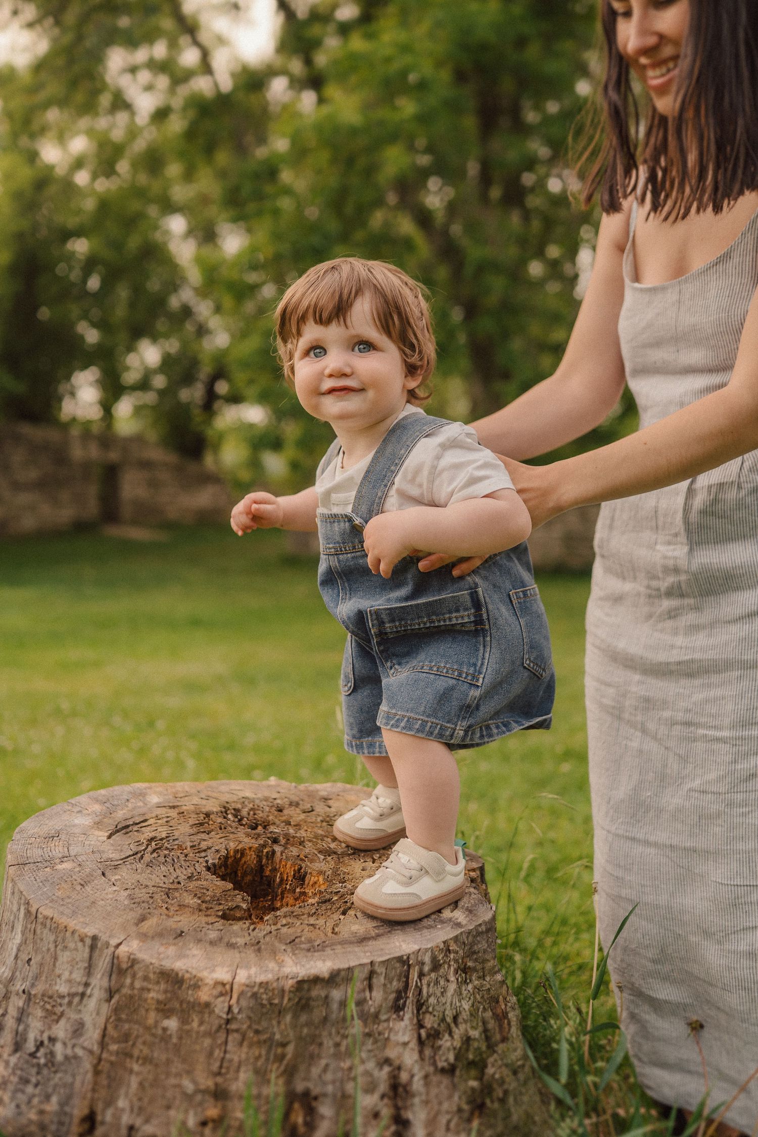 A small child in denim overalls stands on a tree stump while being supported in a sunny garden setting.
