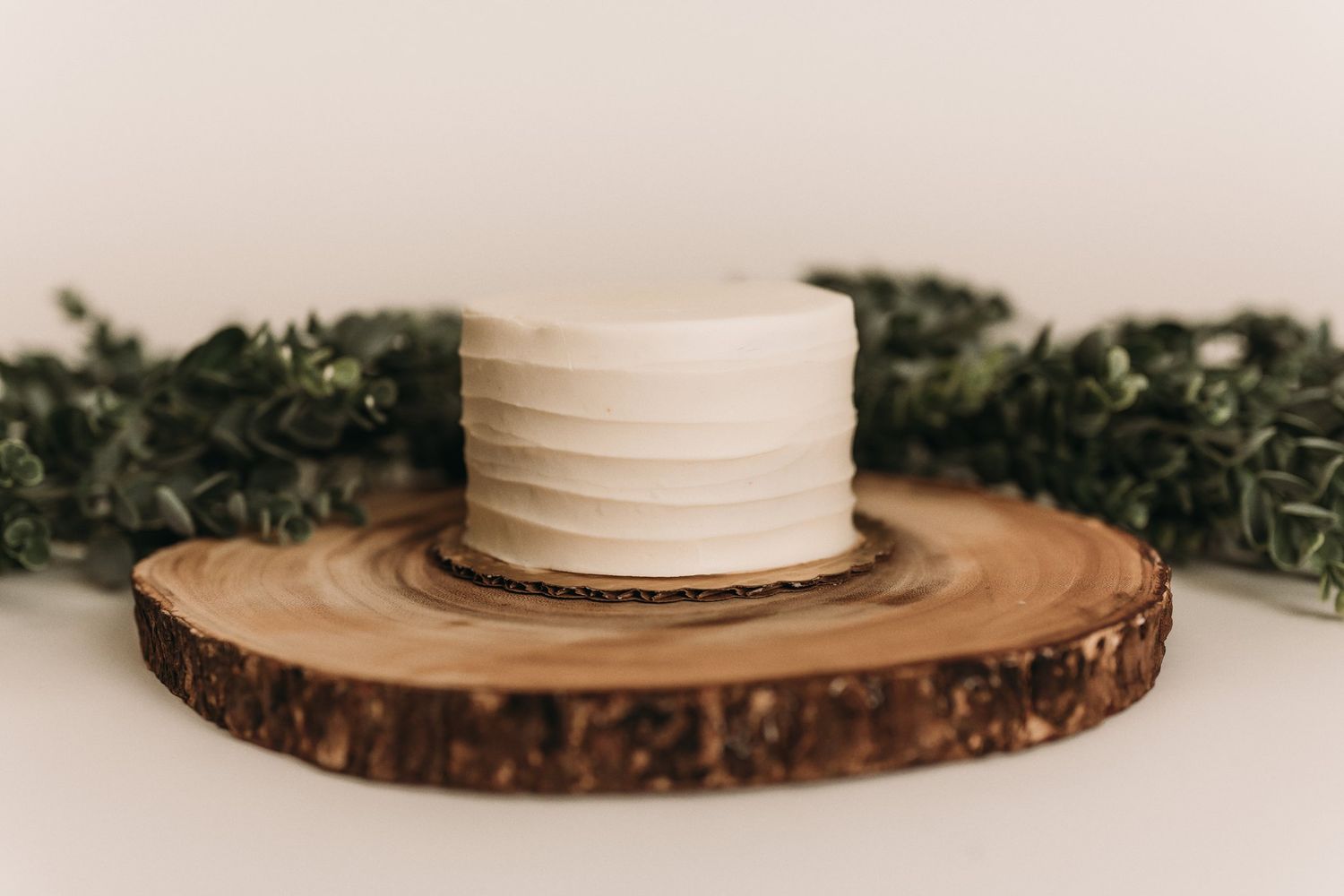 Simple white cake displayed on rustic wood slice with greenery accents.
