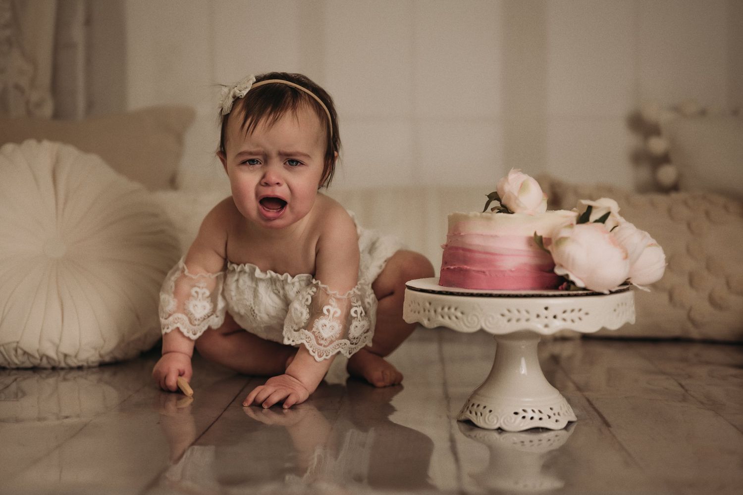 Baby in white lace outfit making expressive face next to pink cake on decorative stand.