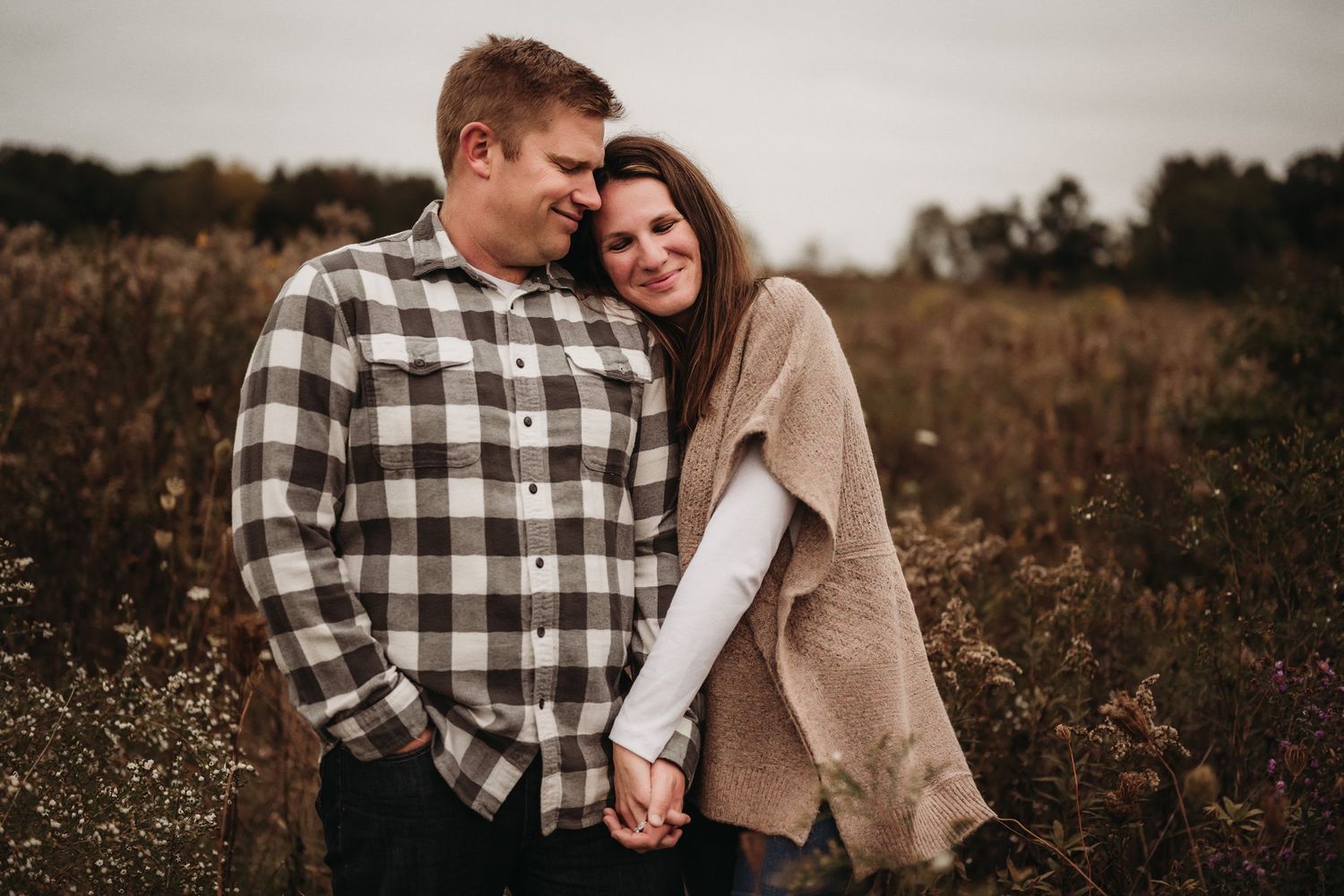 A couple in casual fall attire embraces in a rustic field setting.