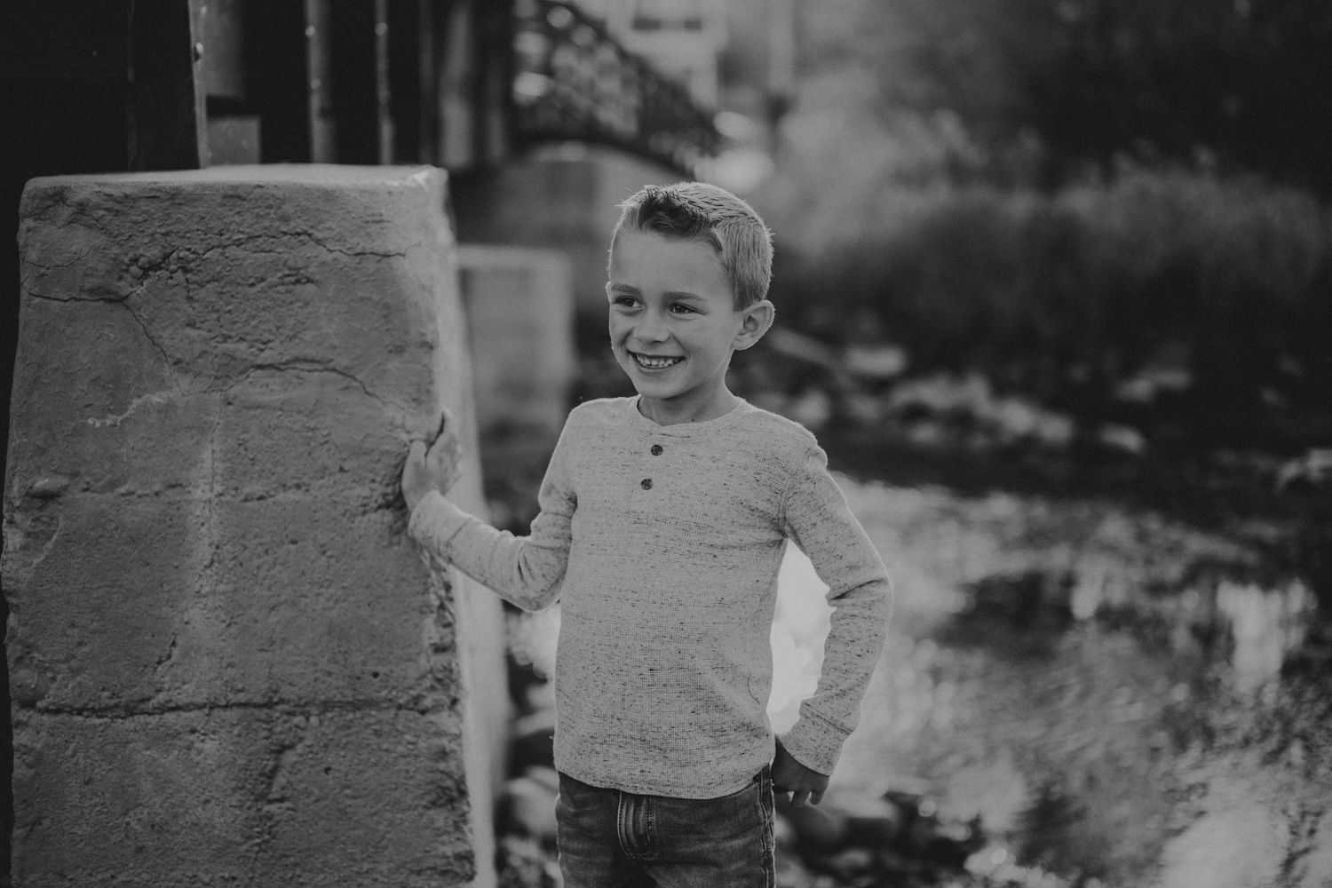 A black and white portrait of a young child in a henley shirt posing next to a stone pillar.