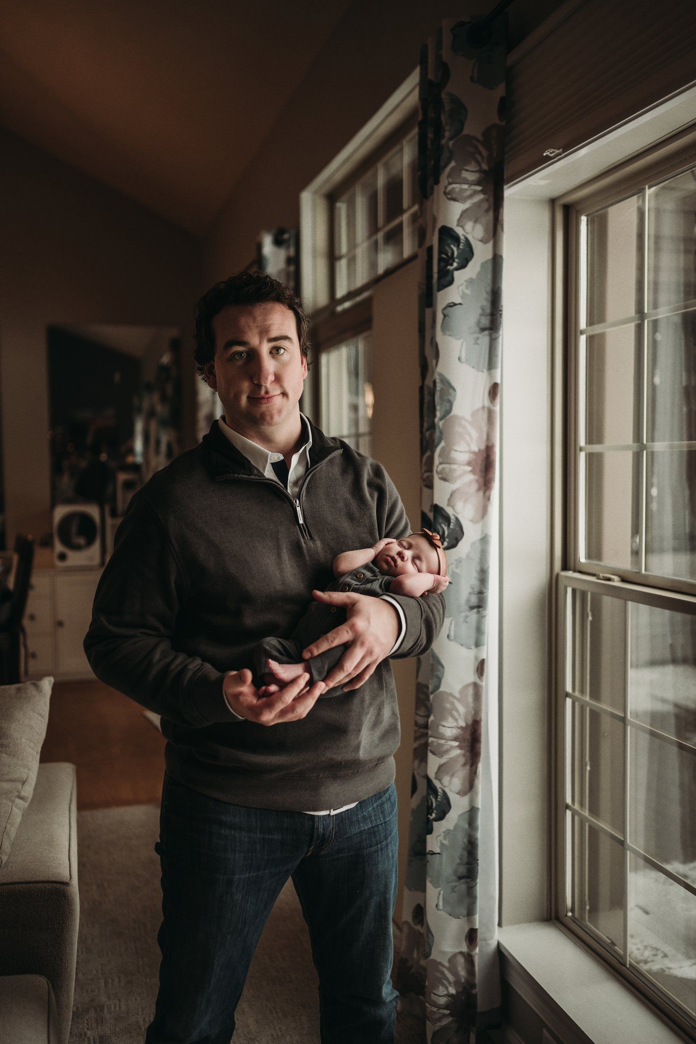 Adult standing by window holding newborn baby in casual home setting.