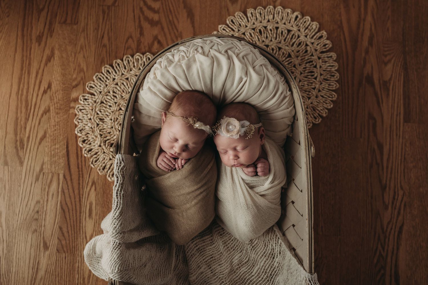 Twin babies swaddled in neutral wraps lying in a decorative bassinet on hardwood floor.
