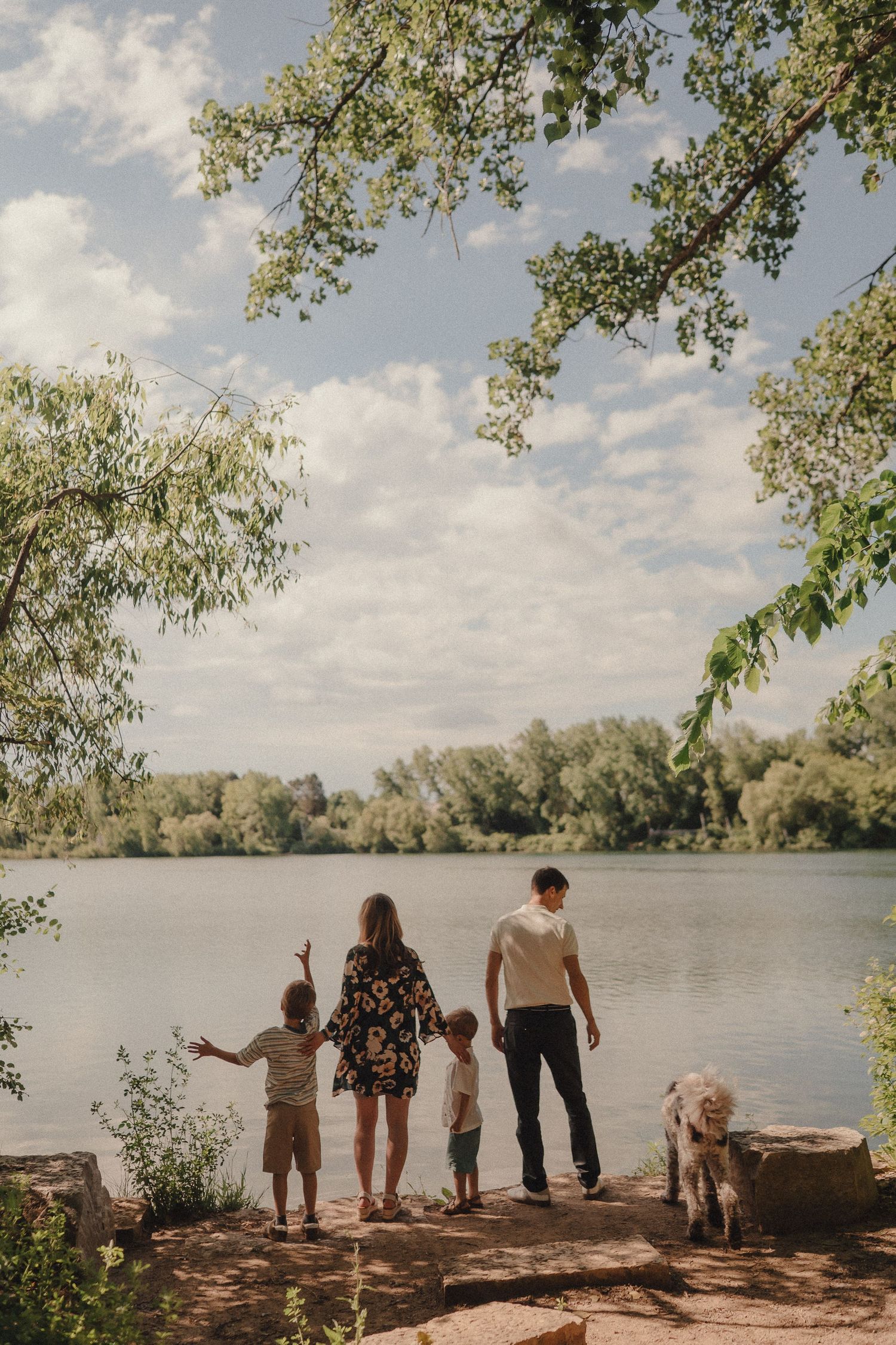 A family with two children and a dog stands by a serene lake framed by overhanging trees.
